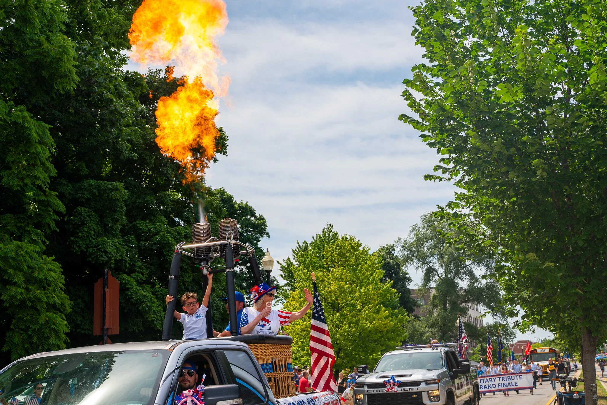 A parade vehicle with a hot air balloon and patriotic decorations, including American flags, participating in a patriotic celebration on a tree-lined street filled with spectators.