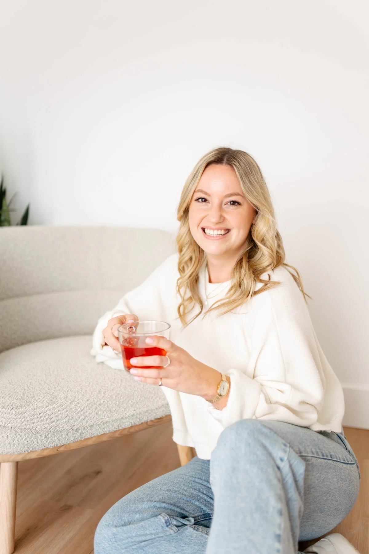 Smiling blonde woman in cream sweater holding herbal tea on a sofa at The Wellness Hub in Stoney Creek, Ontario