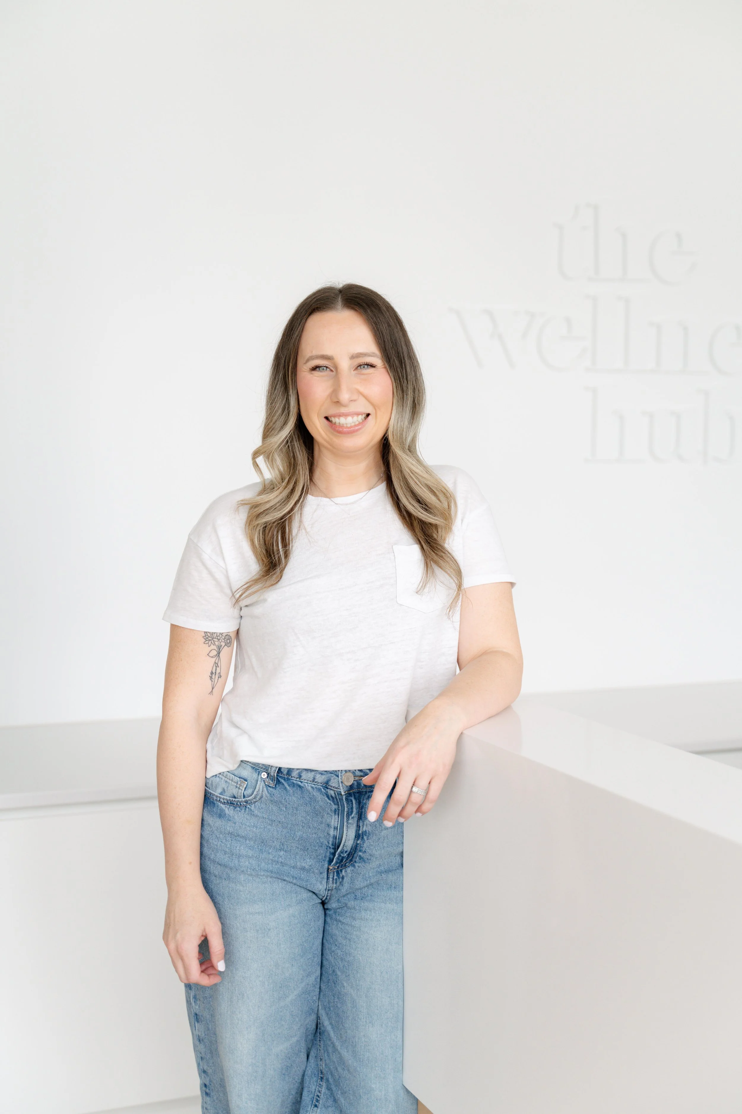 A smiling woman with wavy blonde hair wearing a white t-shirt and blue jeans standing in front of a white wall with faint text, resting one arm on a white counter.