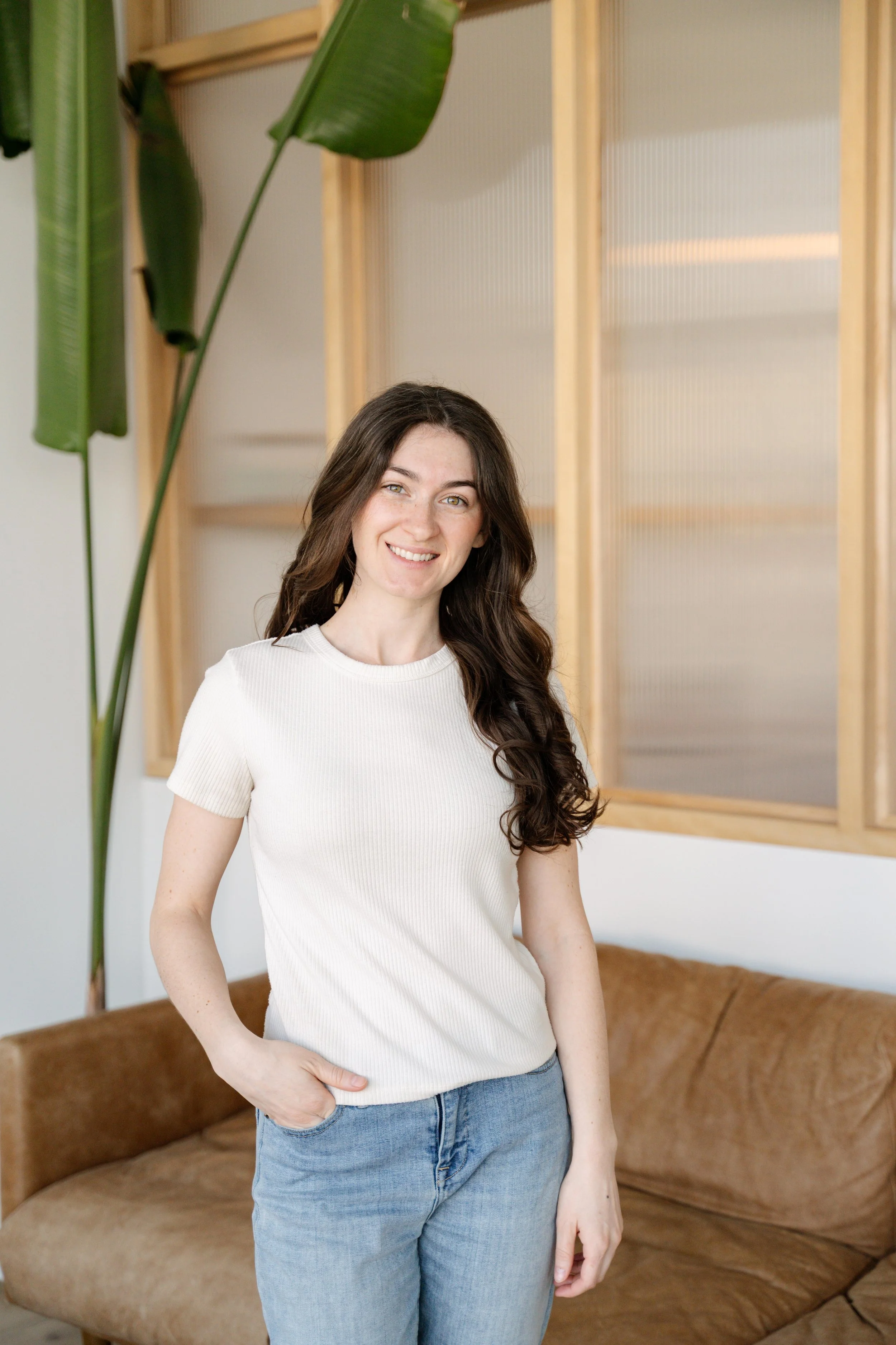 A woman with long brown hair wearing a white T-shirt and blue jeans standing indoors in front of a brown couch and wooden window frame with frosted glass. She is smiling and has one hand in her pocket.
