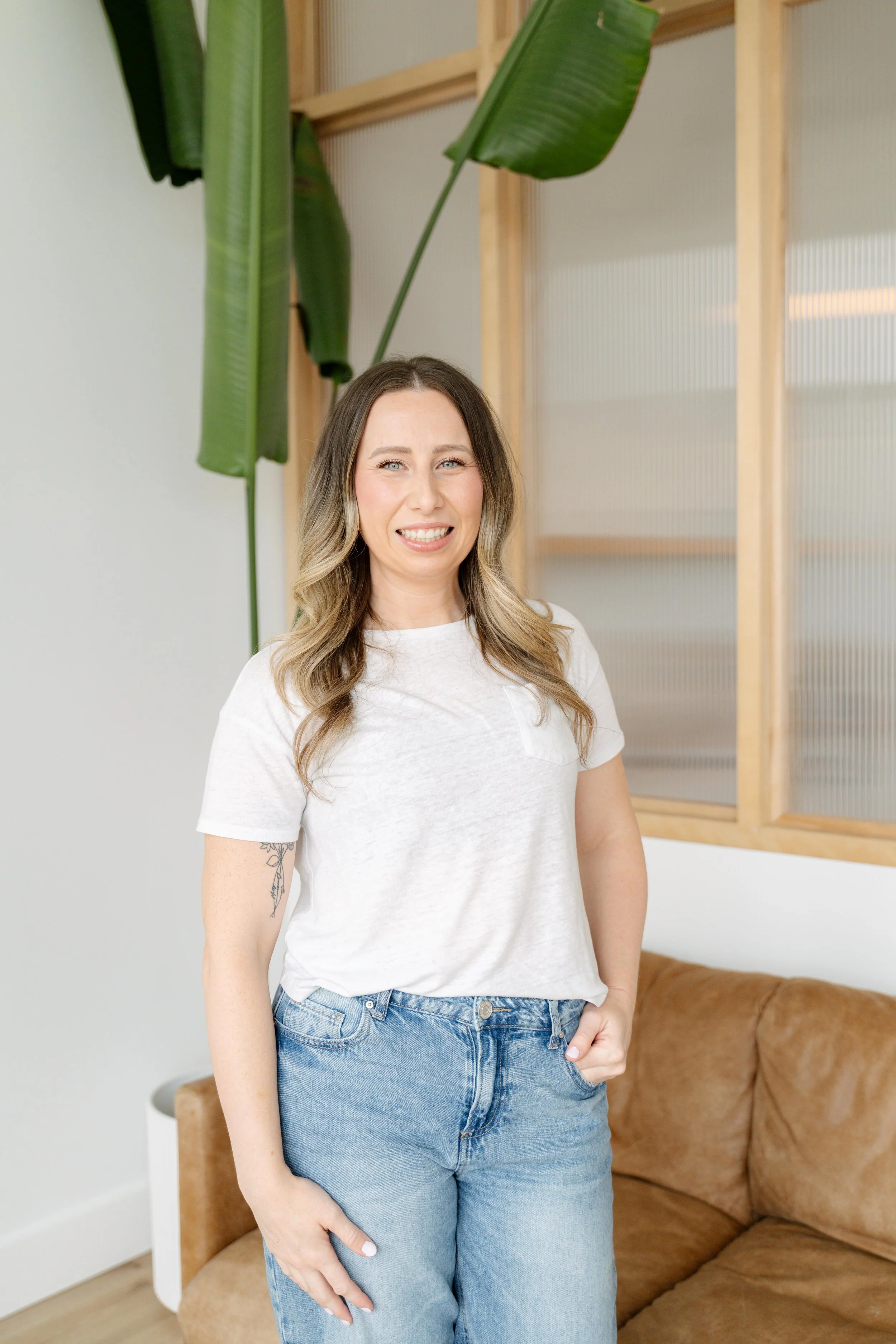 A woman standing indoors, wearing a white t-shirt and blue jeans. She has long, wavy hair and a tattoo on her left arm. She is smiling slightly and has her right hand in her pocket. In the background, there is a brown leather couch, a white side table, a large green plant, and wooden-framed frosted glass windows.