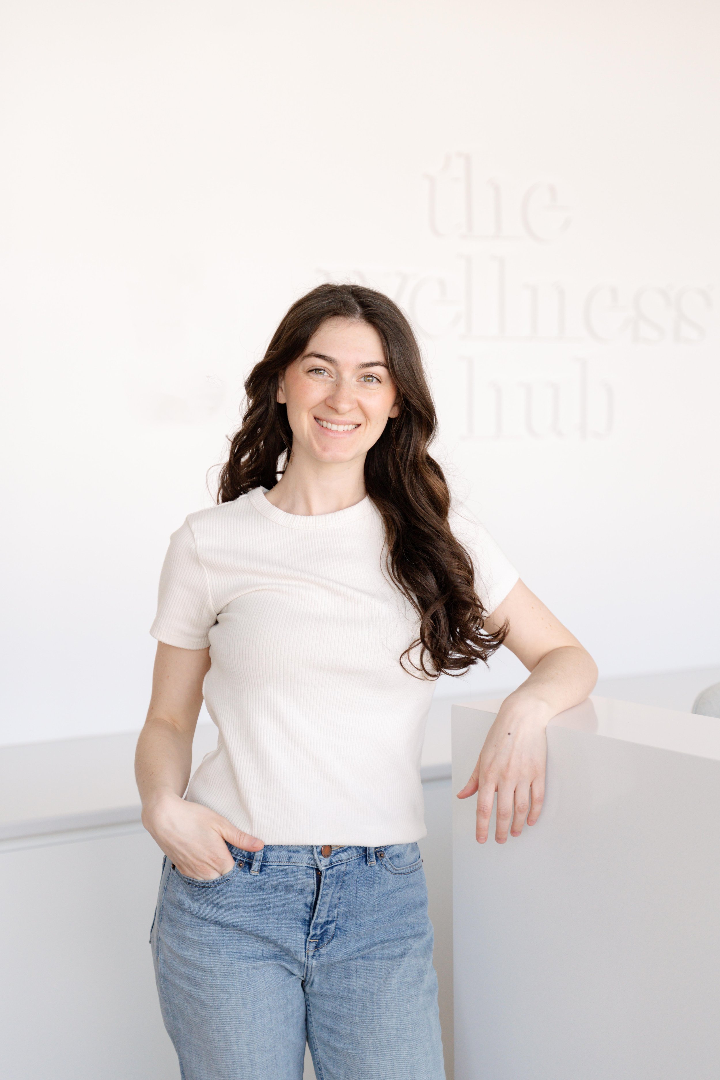 A young woman with long wavy brown hair smiling, standing against a light background with faint text.