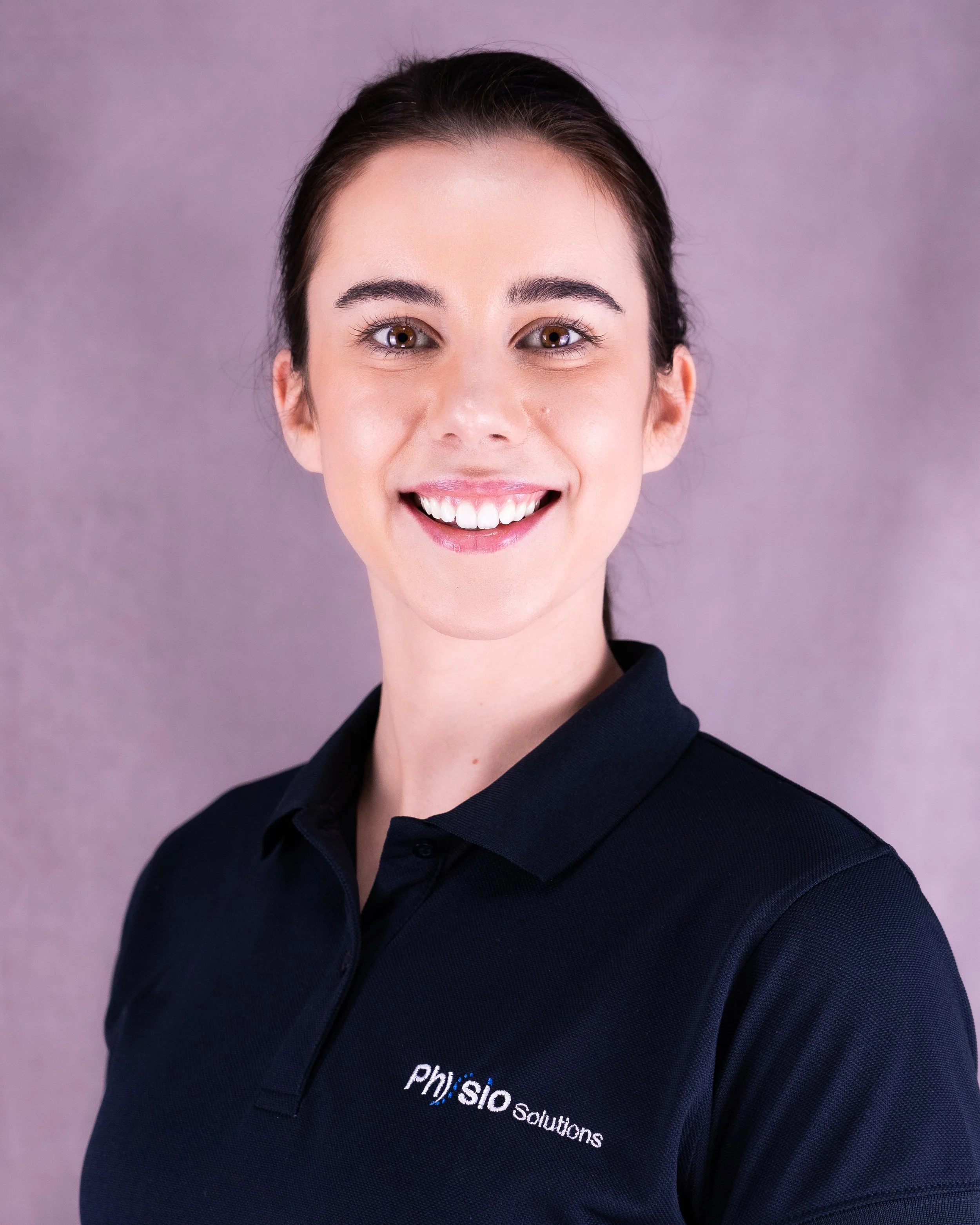 Smiling young woman wearing a black polo shirt with 'Physio Solutions' logo, posing against a purple background.