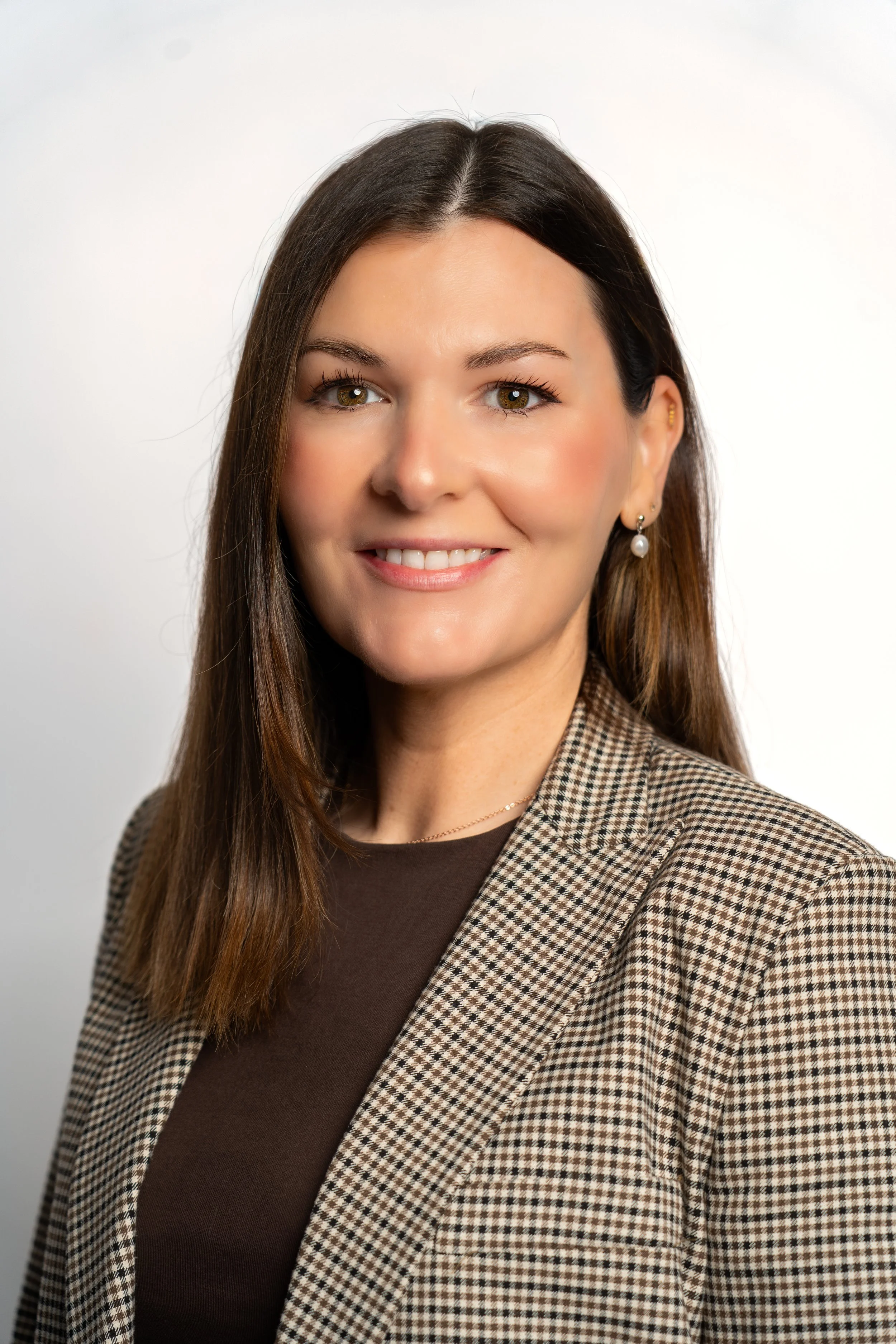 Professional woman with brown hair wearing a checkered blazer and earrings, smiling against a white background.