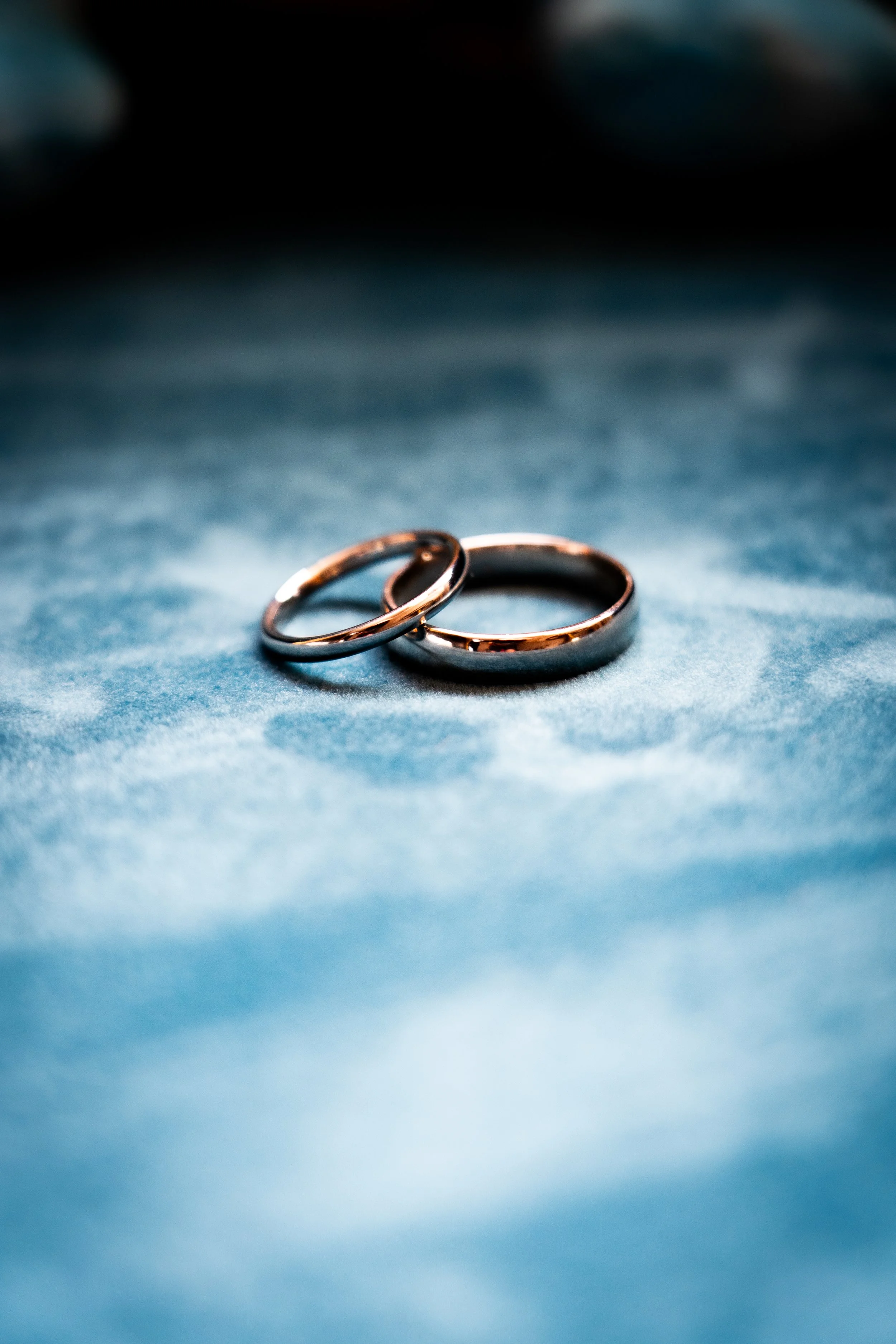 Two wedding rings resting on a soft, velvety blue fabric surface, with a dark background.