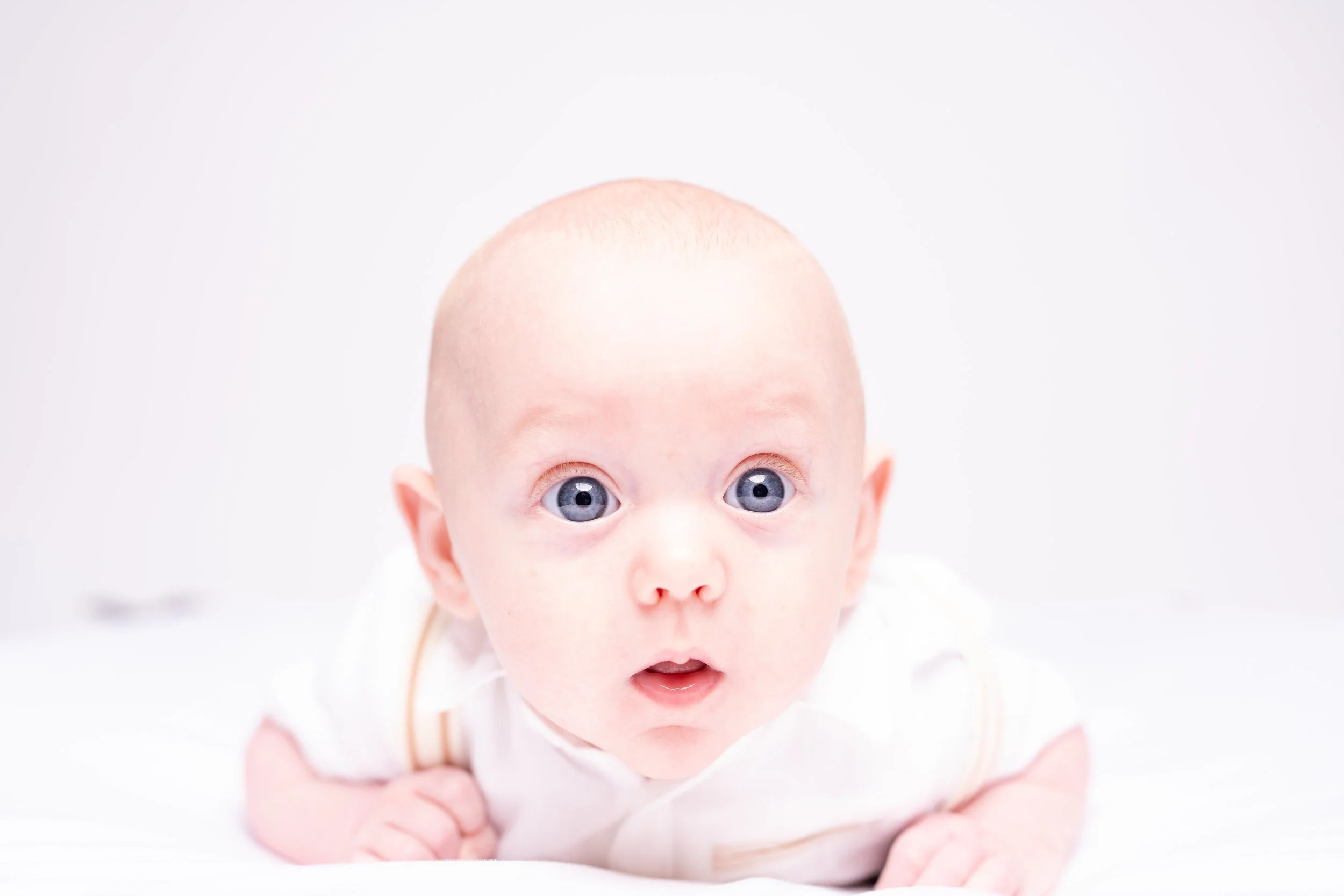 A baby with blue eyes and a bald head lies on its stomach on a white surface, looking directly at the camera with a surprised expression.