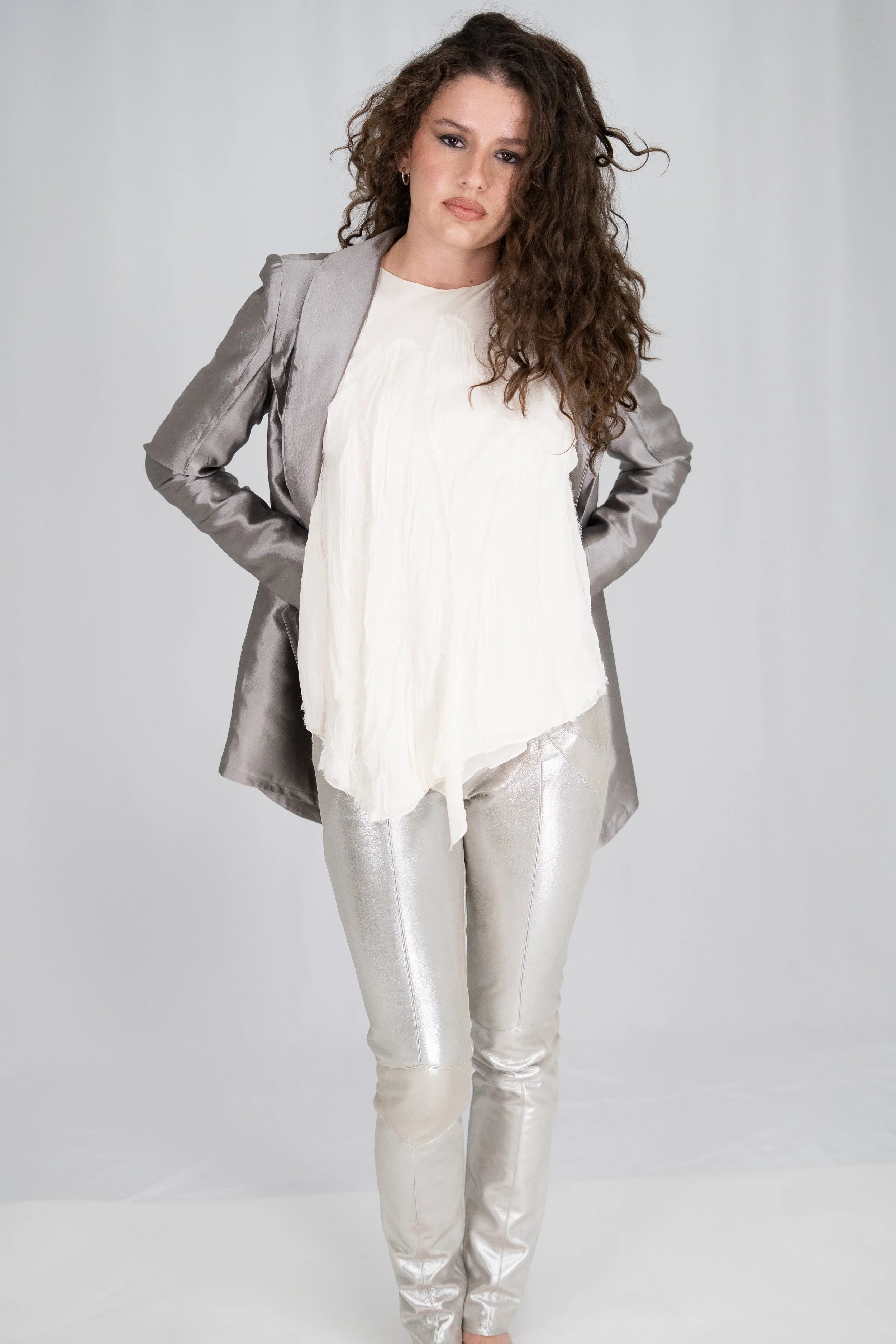 Woman with curly hair wearing a gray blazer, white blouse, and shiny silver pants standing with hands in pockets against a plain gray background.