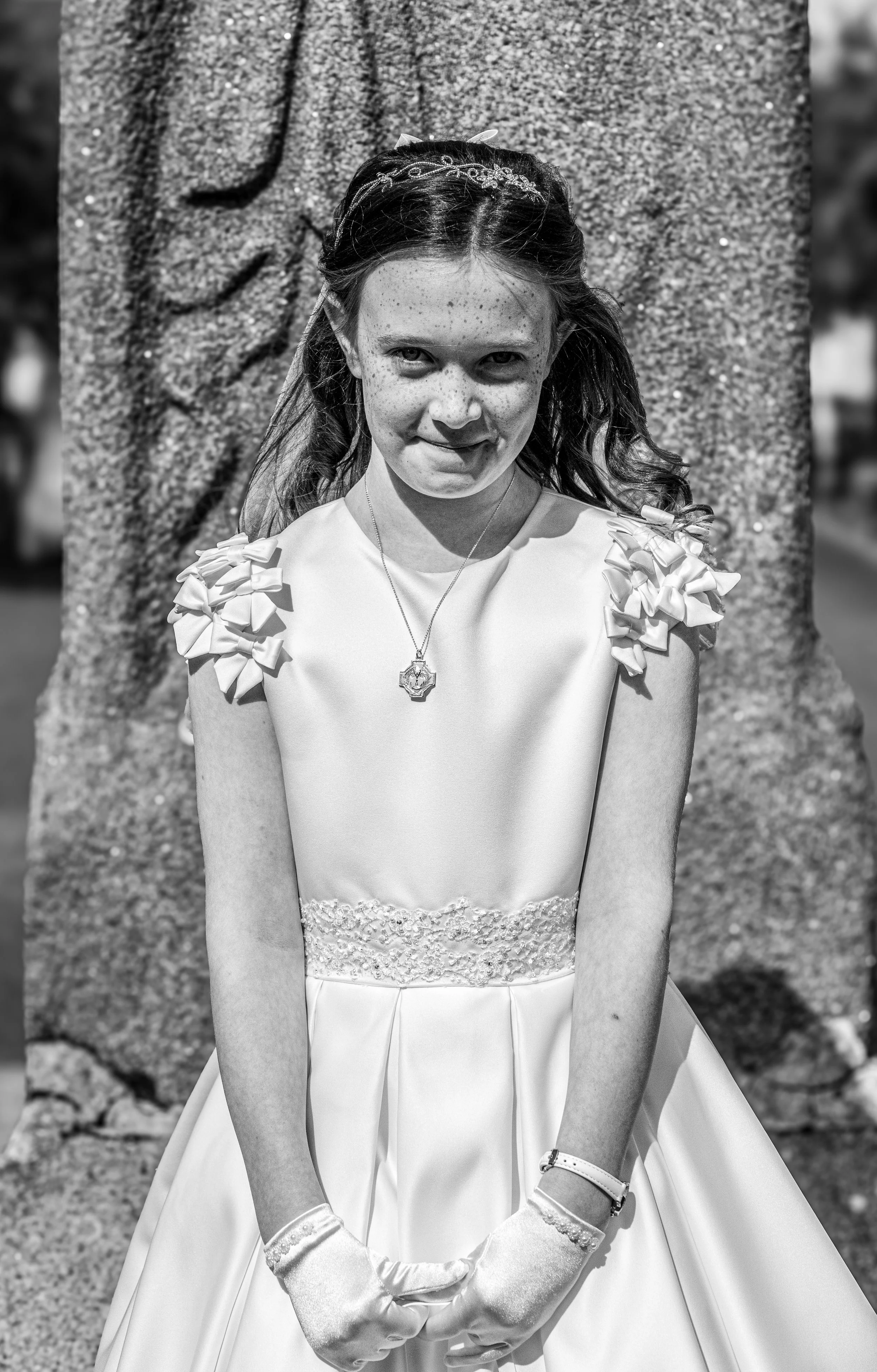 A young girl dressed in a fancy white dress with decorative shoulder bows, wearing gloves, a necklace, and a tiara, standing outdoors in front of a textured rock.