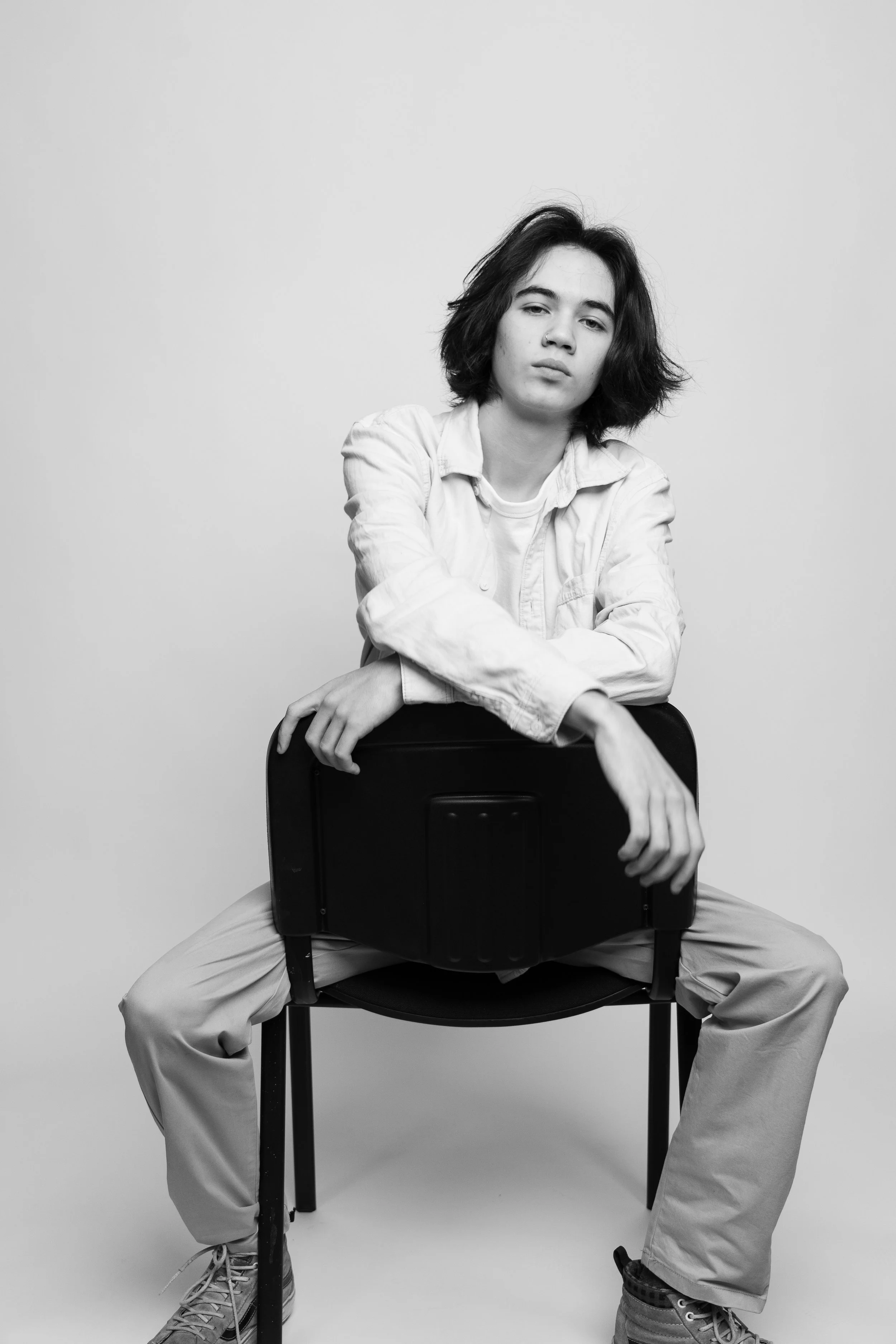 Black and white photo of a young woman with shoulder-length hair, sitting on a chair with a suitcase, wearing a white shirt and light-colored pants, against a plain background.