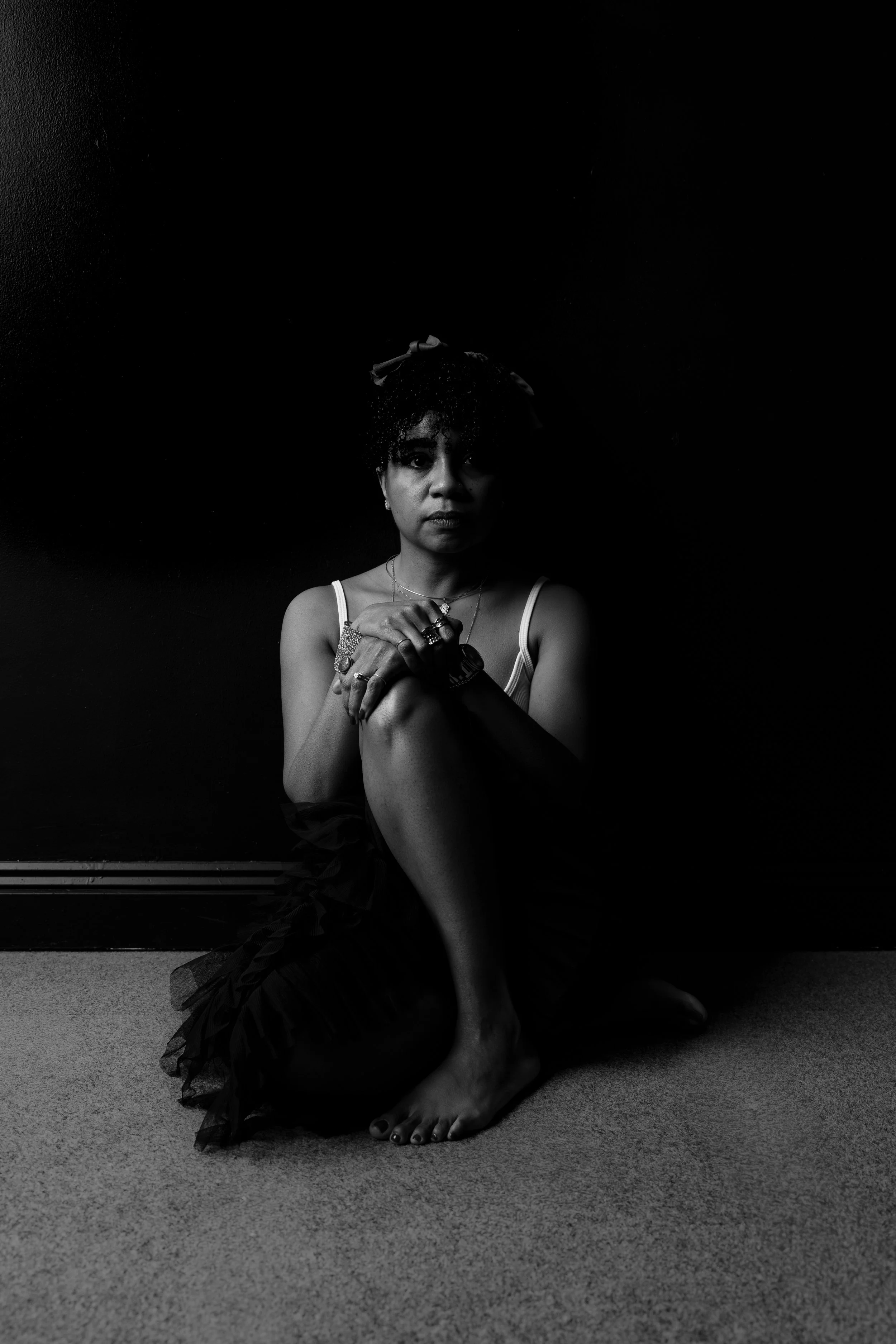 A black and white portrait of a woman sitting on the floor with one leg bent and the other crossed over it, against a dark background. She is looking directly at the camera, wearing a sleeveless top and layered jewelry.
