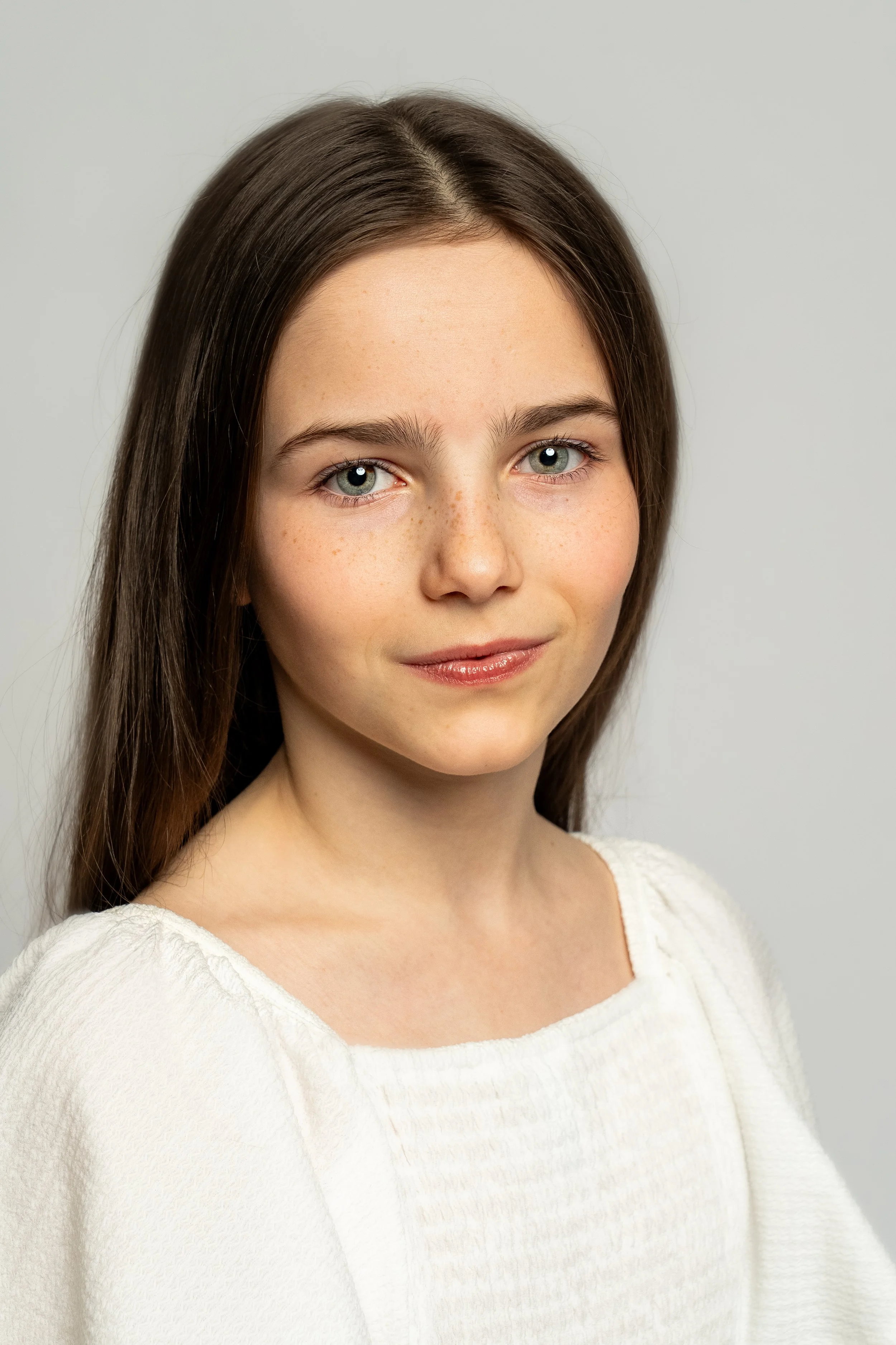 A young woman with long brown hair, blue eyes, and freckles wearing a white top, looking at the camera against a plain light background.