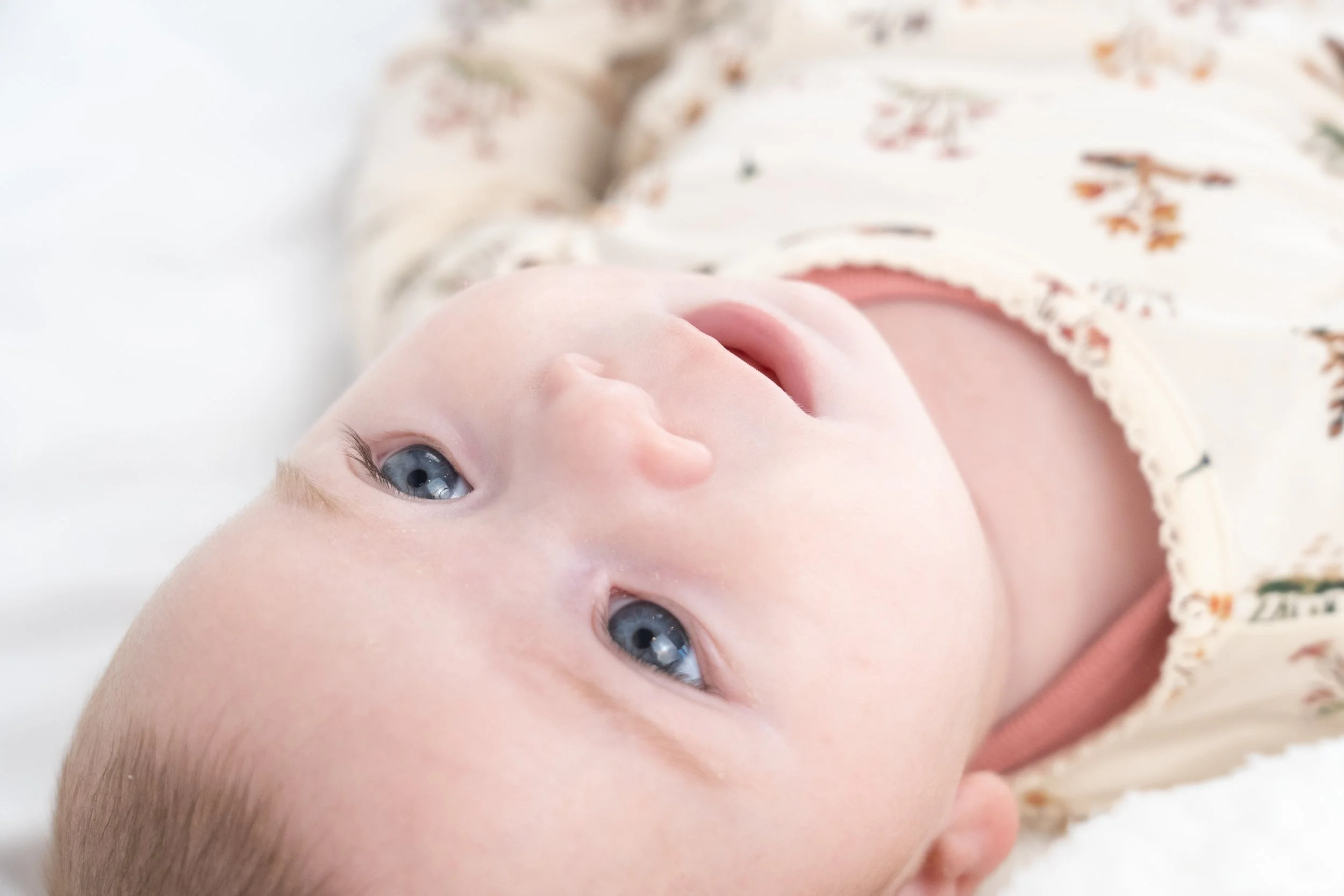 Close-up of a baby lying on a white surface, wearing a floral print shirt, with blue eyes and light skin.