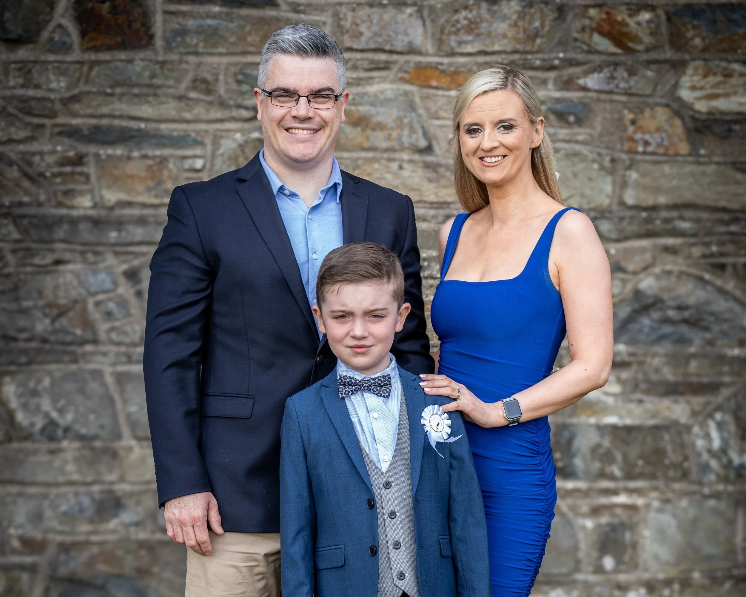 A family portrait of a man, woman, and boy standing outdoors in front of a stone wall, dressed formally, smiling at the camera.