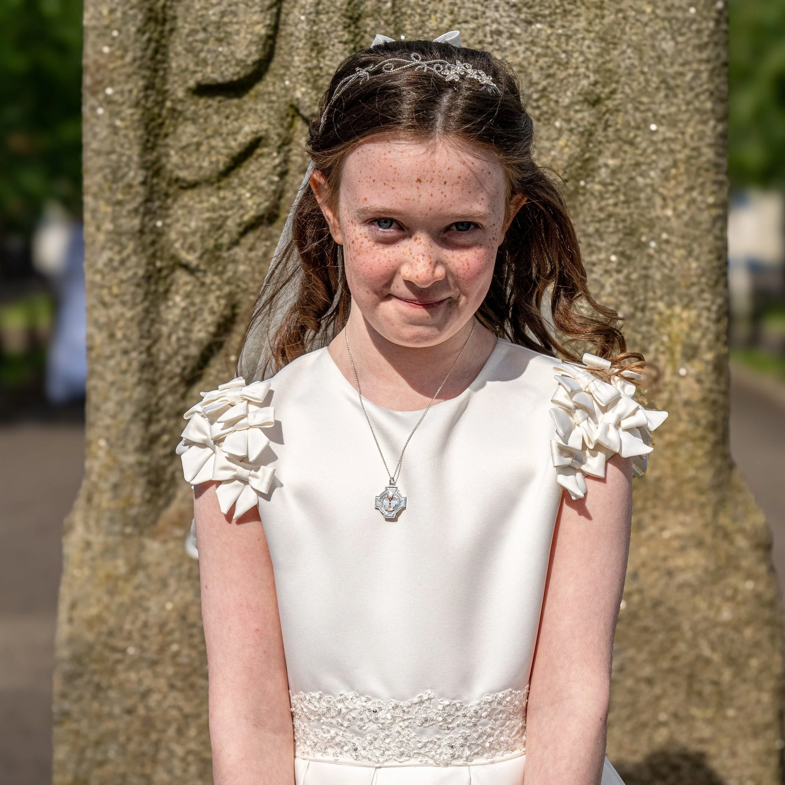 Young girl with freckles wearing a white dress with ruffled sleeves, a necklace, and a headband, standing in front of a tree outdoors.