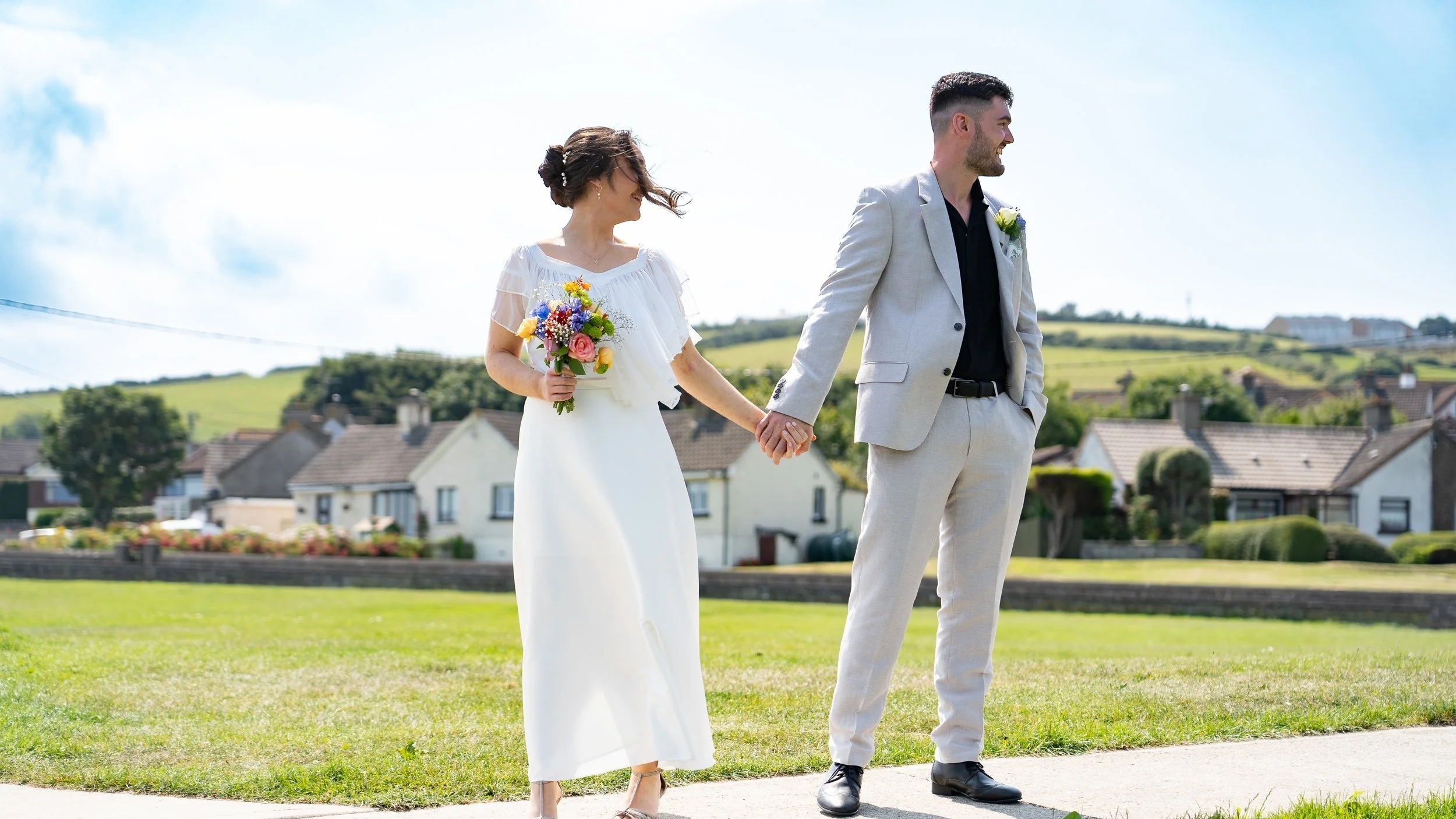 A couple dressed in wedding attire holding hands outdoors on a sunny day, with houses and rolling hills in the background.