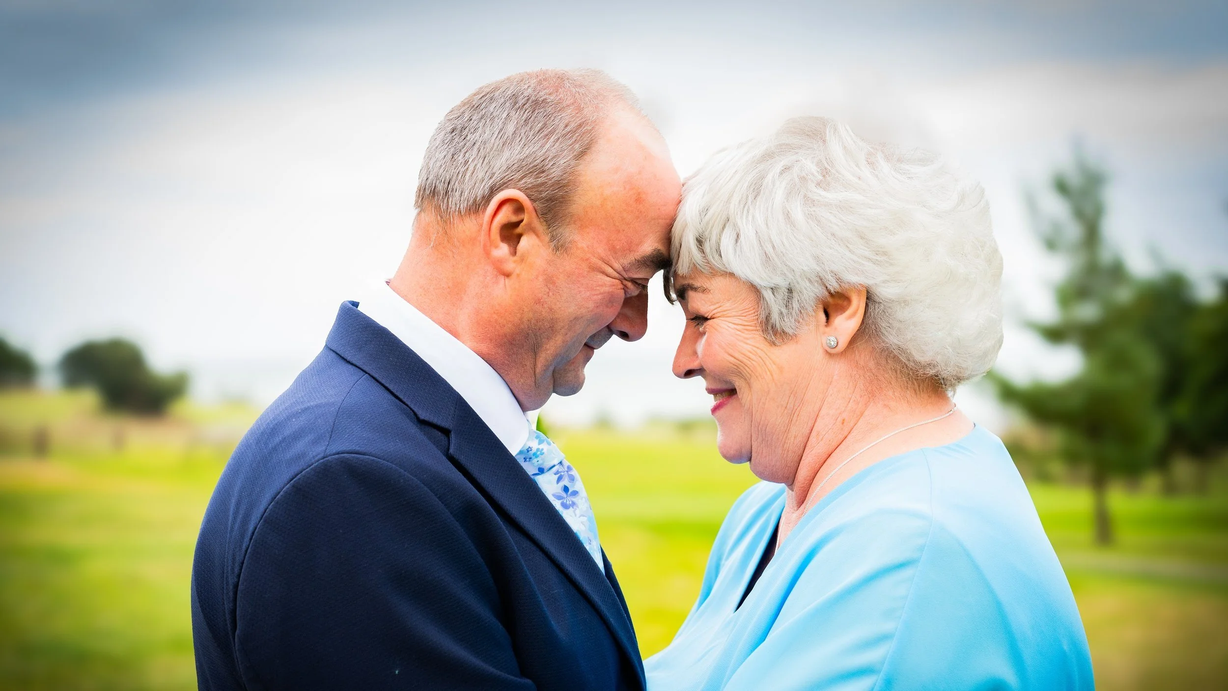 A happy elderly man and woman with foreheads touching, smiling at each other outdoors in a grassy area with trees in the background.