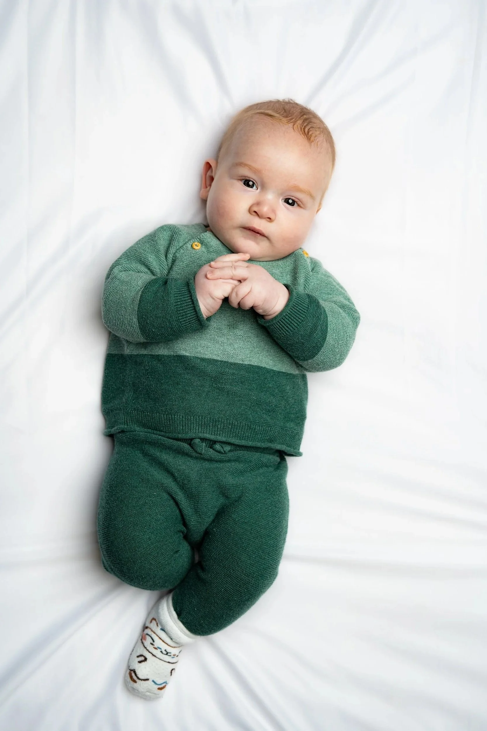 Young child lying on a white sheet, wearing green sweater and pants, with socks featuring a playful pattern.