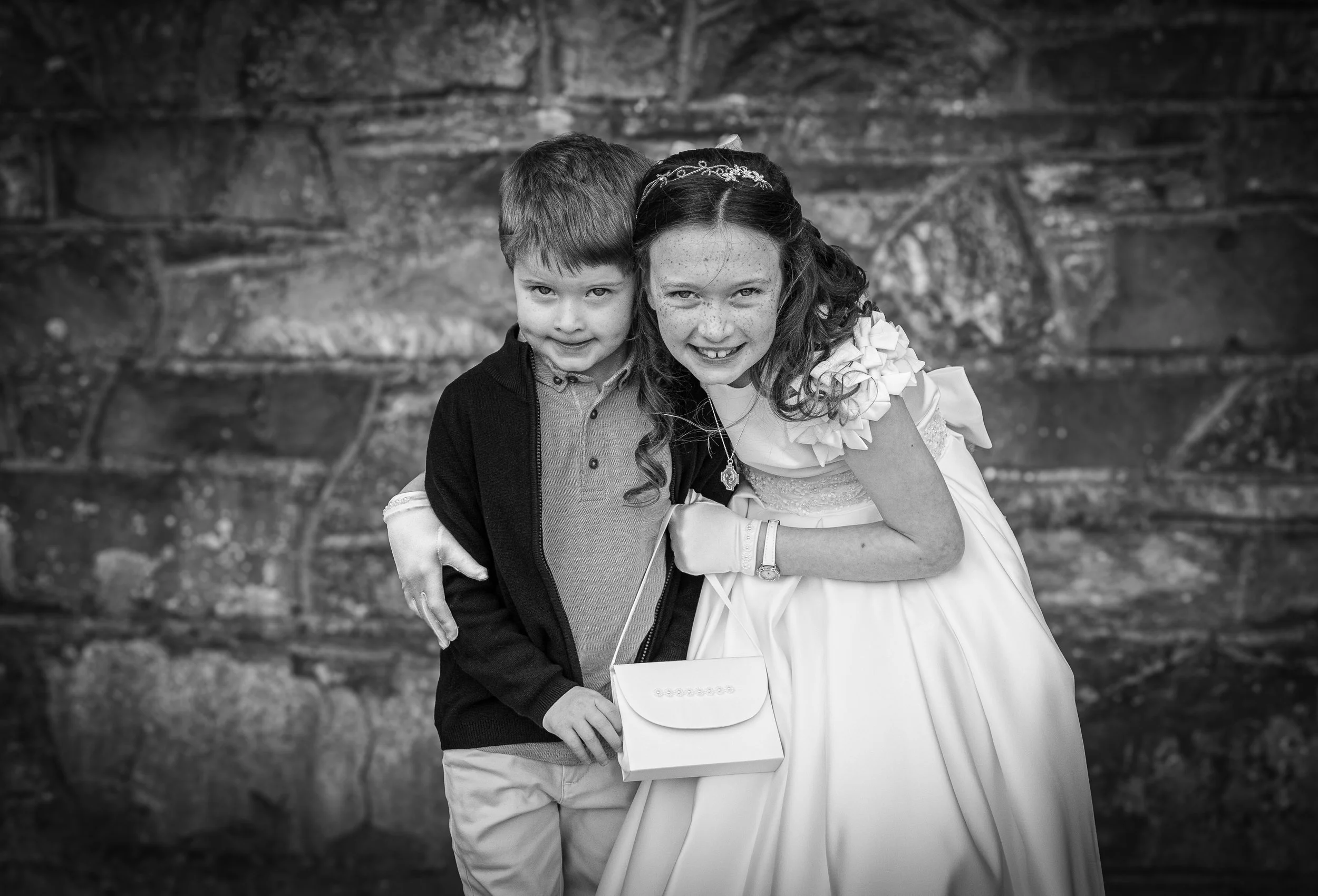 Black and white photo of a young boy in a white shirt and a young girl in a white dress with a headband, smiling at the camera