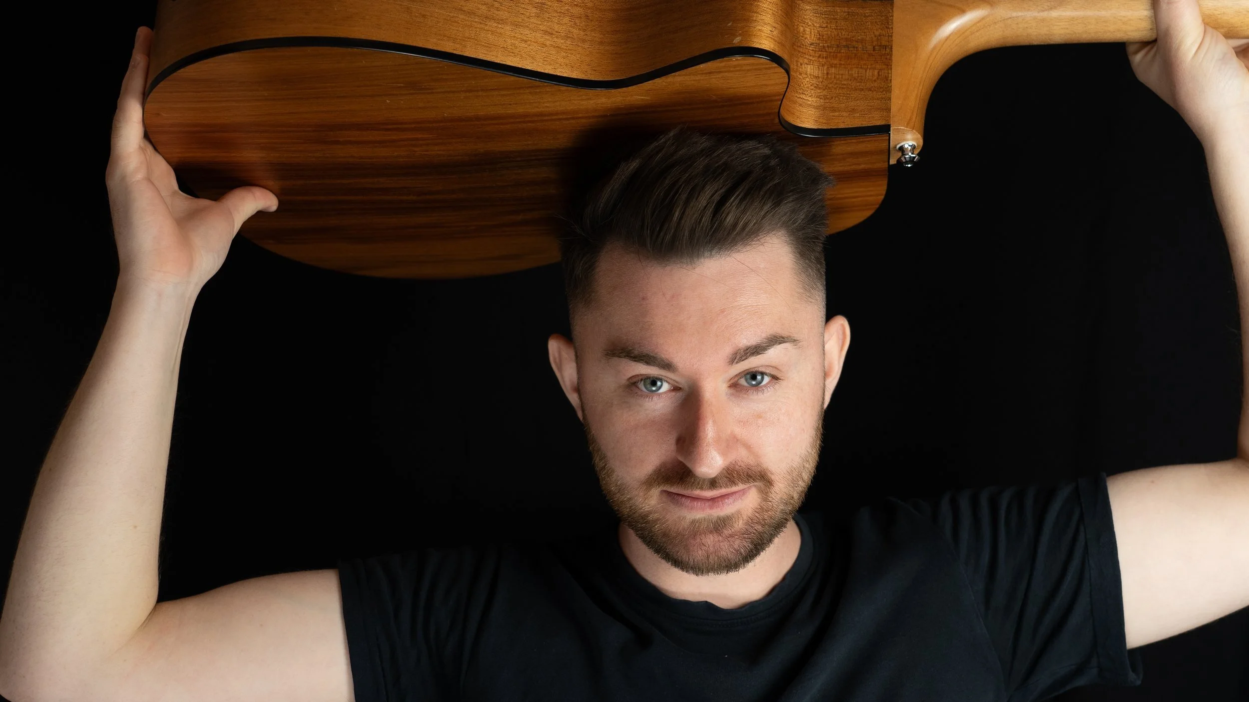 A young man with short brown hair and a beard, lying down on a black surface, holding a wooden musical instrument above his head, looking up at the camera.
