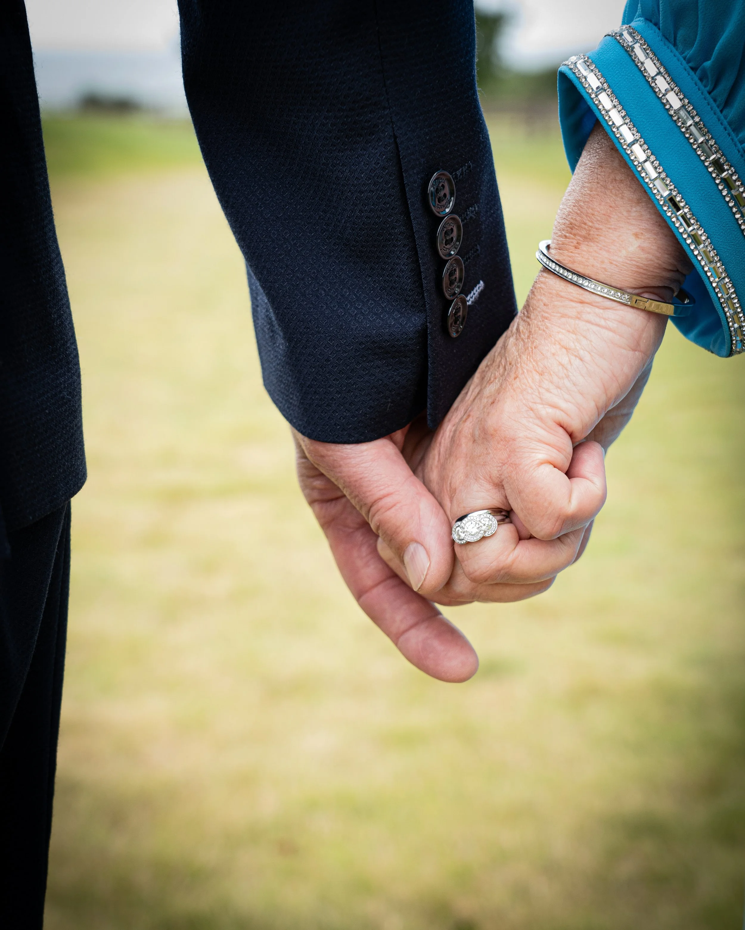 Close-up of a couple holding hands, with the woman wearing a blue dress with decorative trim and a silver bracelet, and both showing rings on their rings fingers, indicating they are engaged or married.