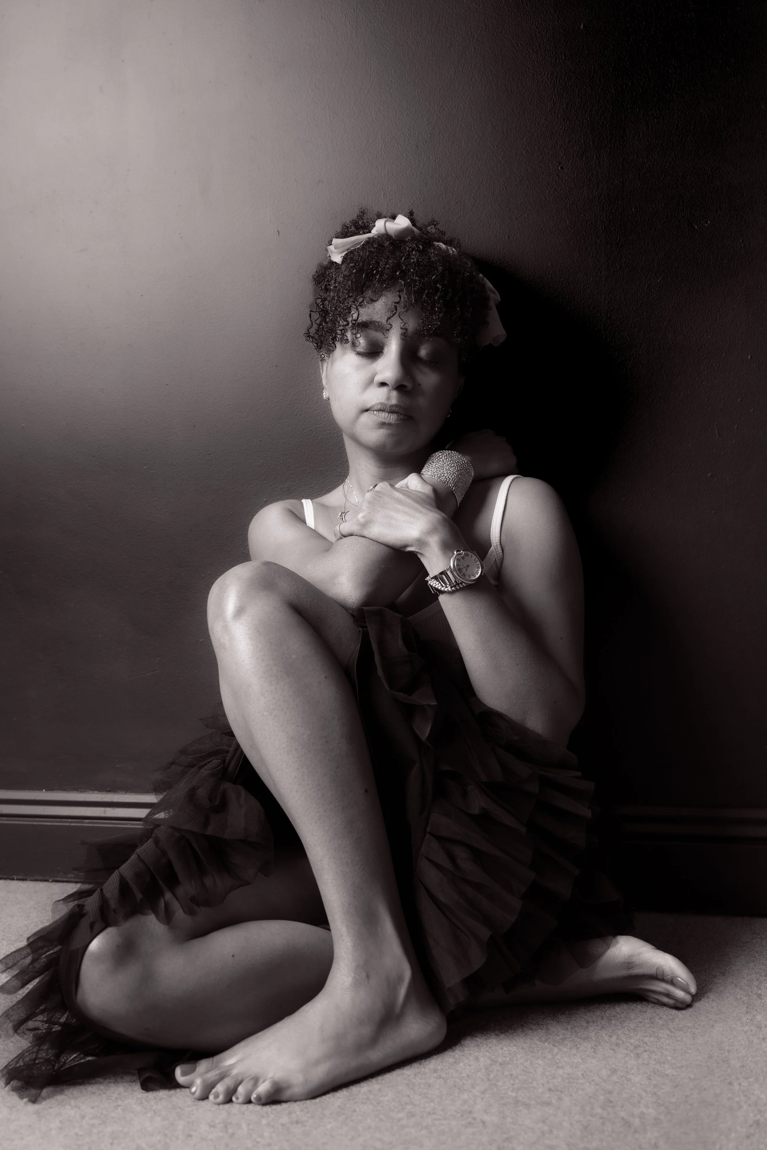 A woman sitting on the floor with her eyes closed, touching her shoulder, against a dark wall background, dressed in a sleeveless top and a ruffled skirt.