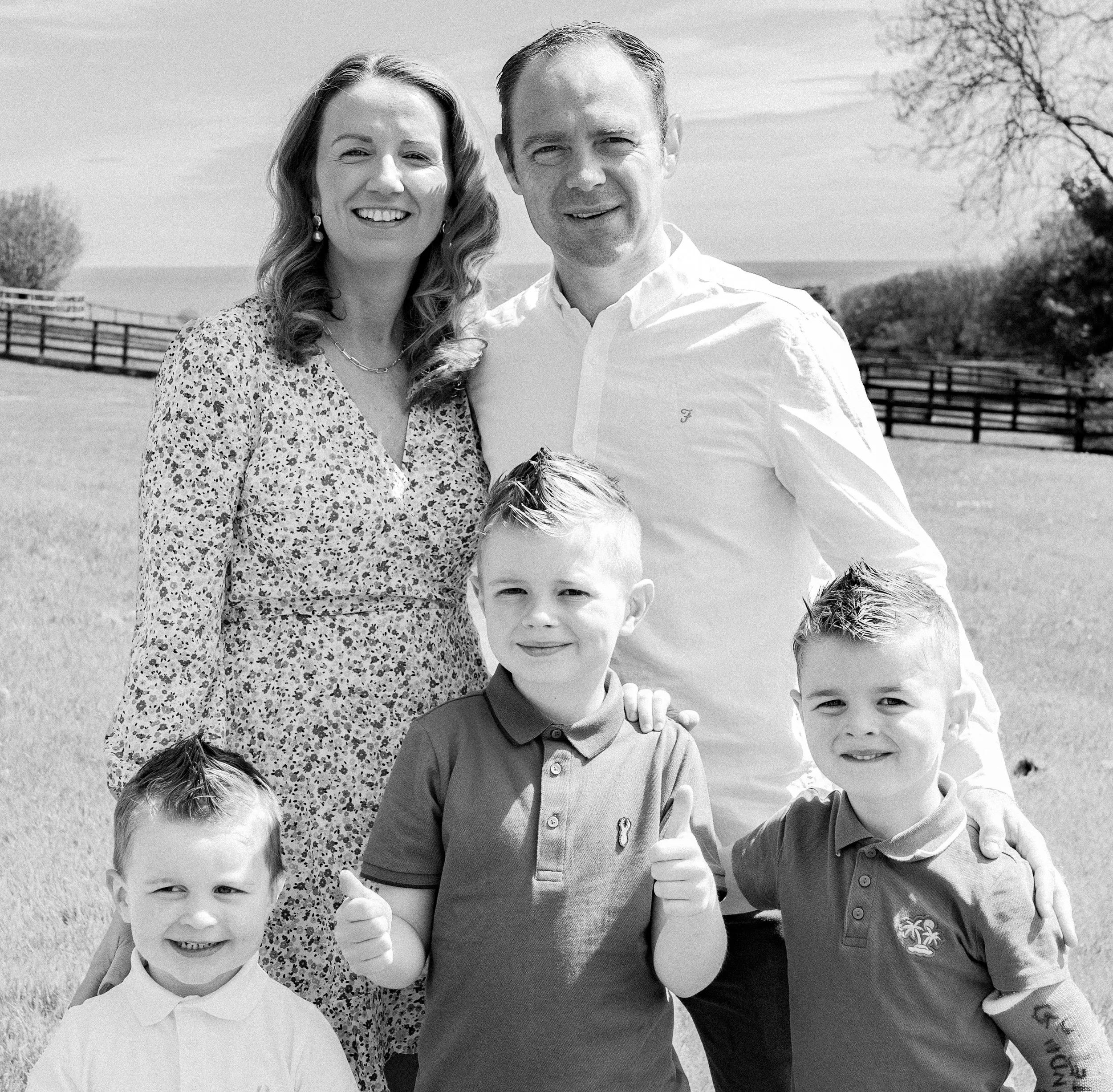 A happy family of five standing outdoors in a field. The adults are a woman with long wavy hair wearing a floral dress, and a man with short hair in a white dress shirt. There are three young boys in front, two giving thumbs-up, all smiling.