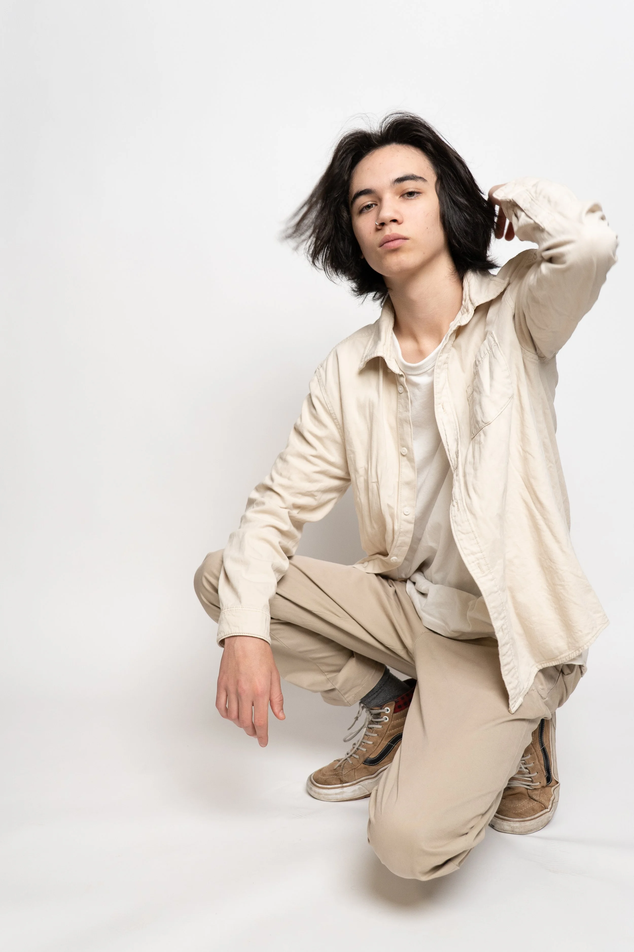 A young person with shoulder-length black hair, wearing a cream-colored button-up shirt and beige pants, crouching with one knee on the ground, against a plain white background.