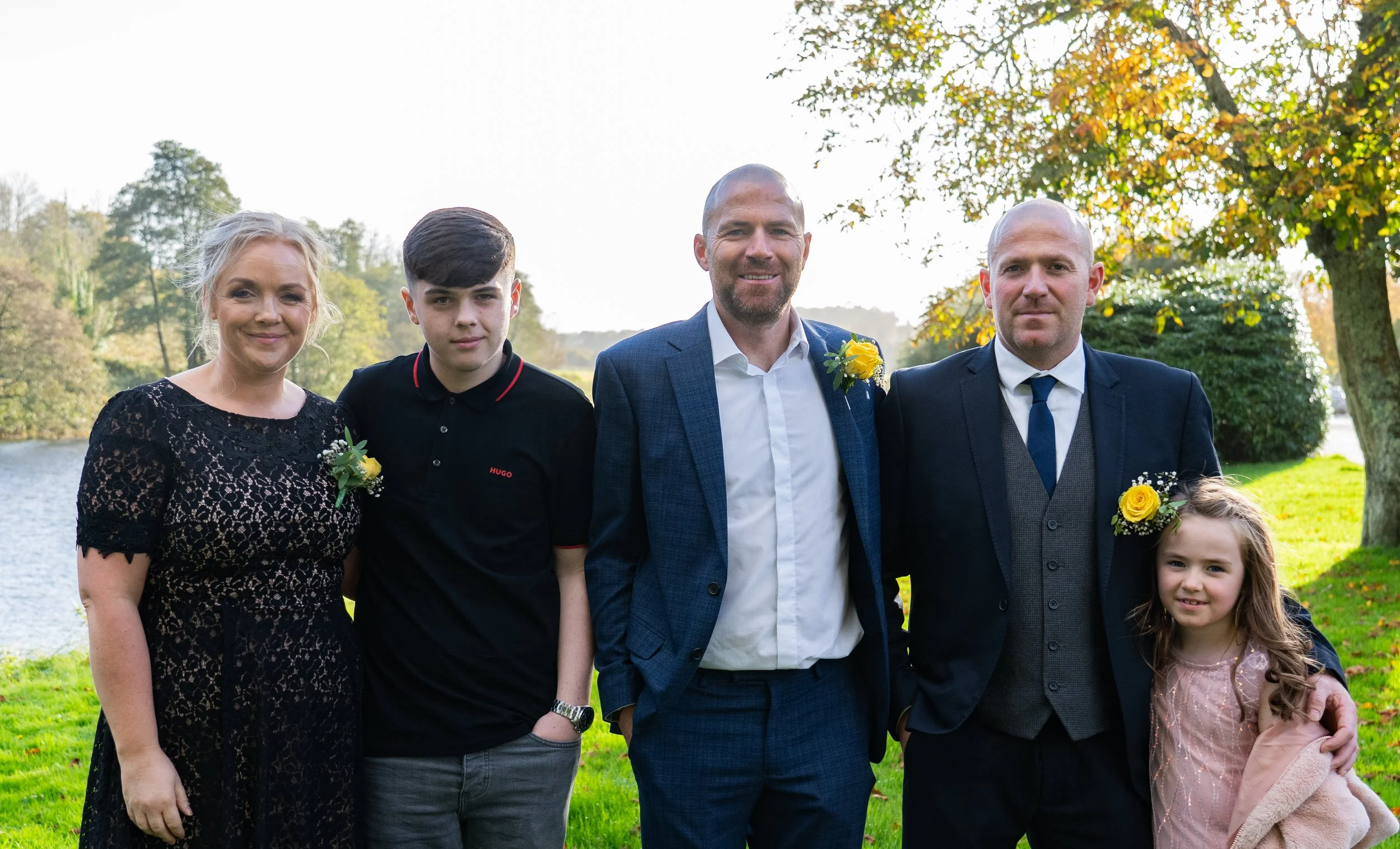 Five people standing outdoors in a park-like setting during daytime, smiling. The group includes two women, two men, and a young girl, all dressed semi-formally with yellow flower boutonnieres and a corsage.