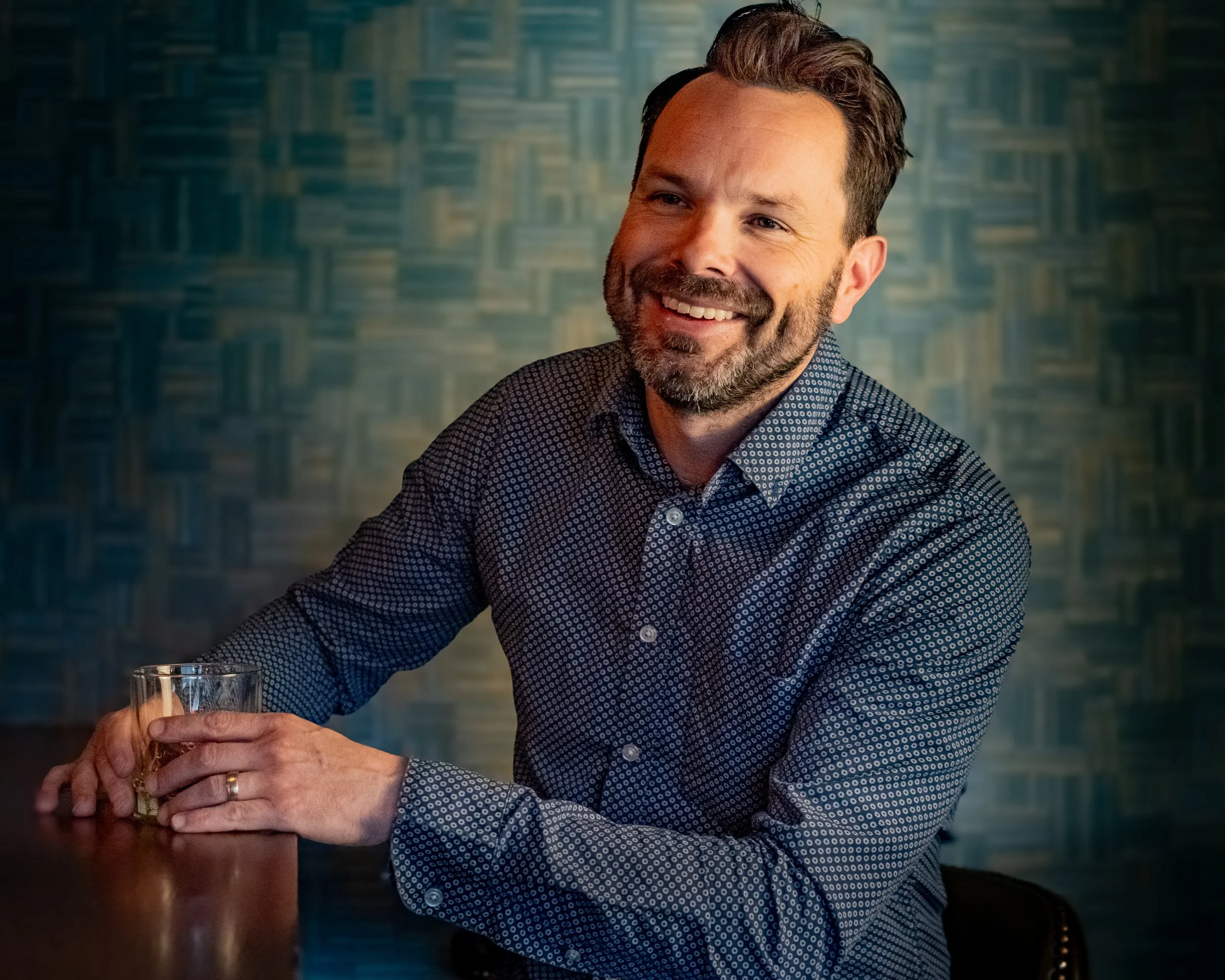A man with dark hair and a beard, wearing a patterned dress shirt, sitting at a wooden table with a glass of drink in his hand, smiling against a patterned blue-green background.