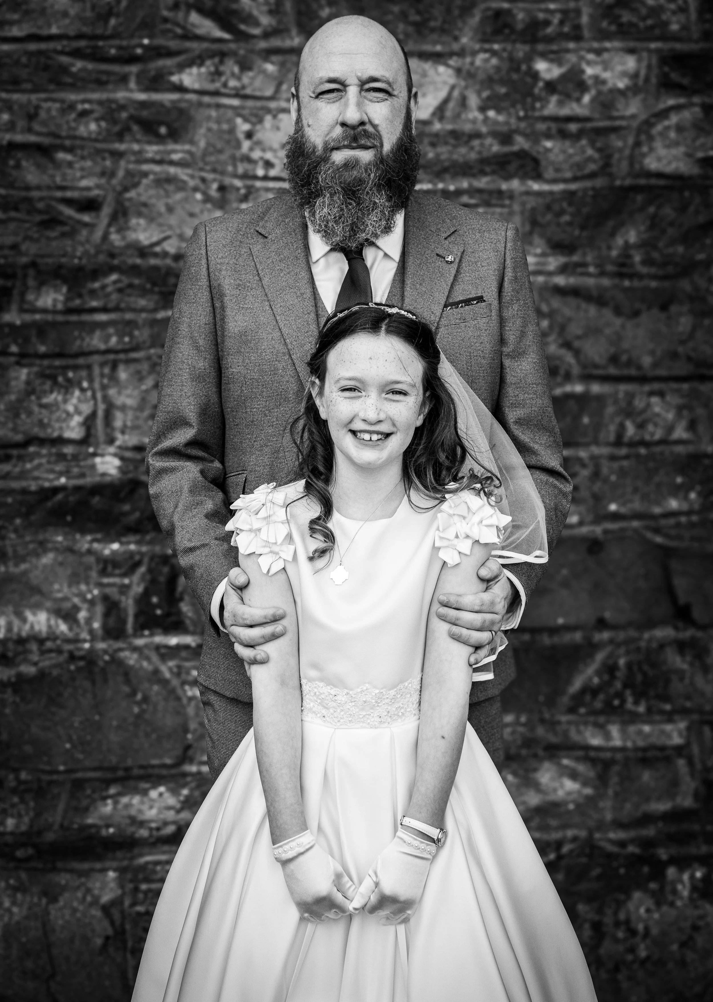 A man and a young girl posing for a wedding photo outdoors, with a stone background. The man is wearing a suit and has a beard, and the girl is in a wedding dress with a veil, smiling.