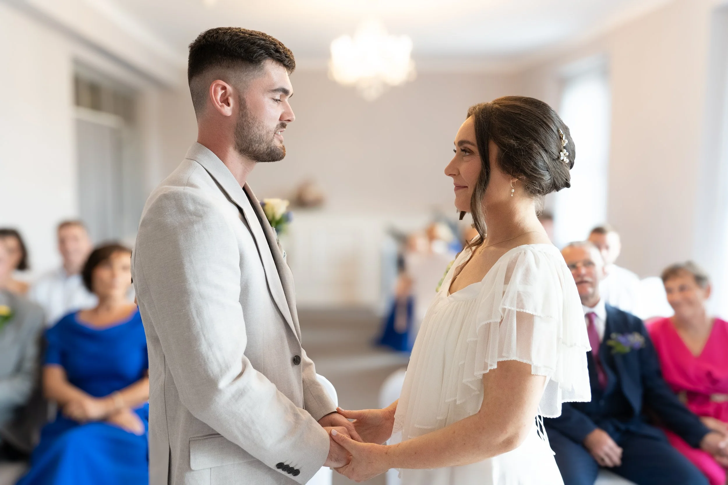 A bride and groom holding hands and looking at each other during their wedding ceremony in a bright room with guests seated in the background.