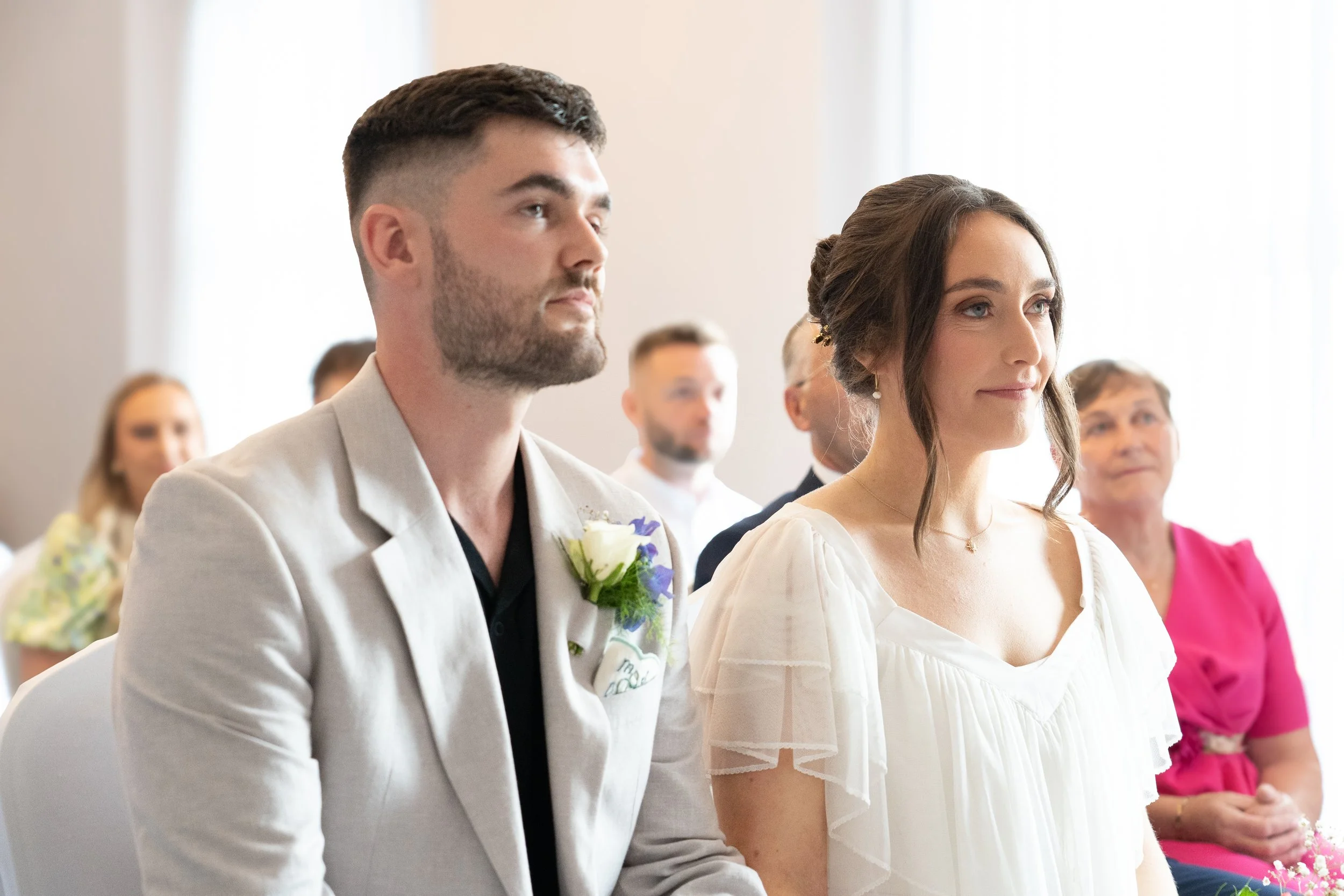 A wedding ceremony with a man in a light-colored suit with a boutonnière, a woman in a white dress, and several guests in the background.