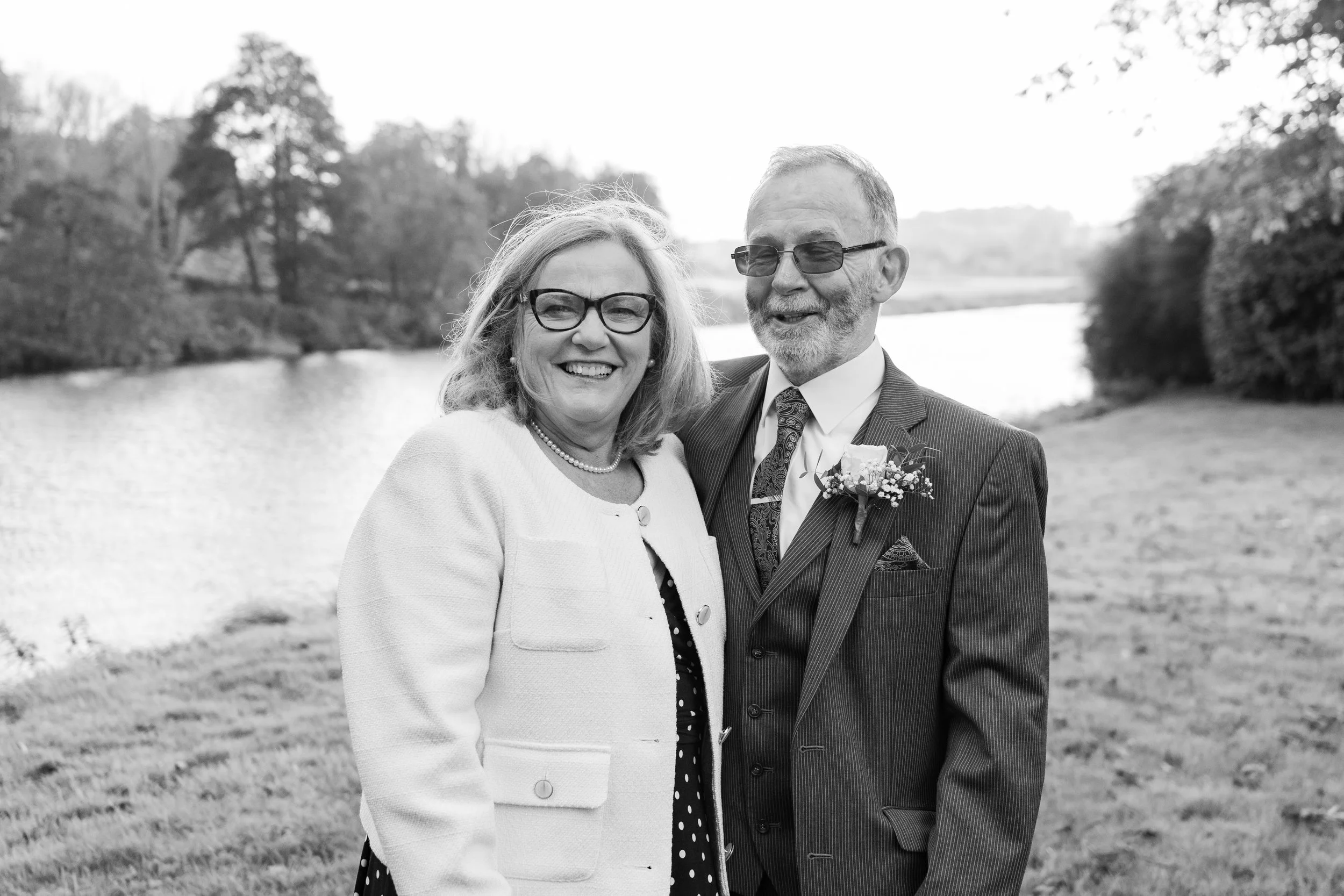 A smiling middle-aged couple in formal attire standing outdoors near a body of water with trees in the background.