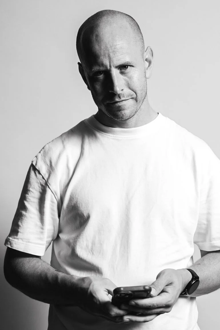 Black and white photo of a bald man wearing a white T-shirt, holding a smartphone, and looking at the camera with a slight smile.