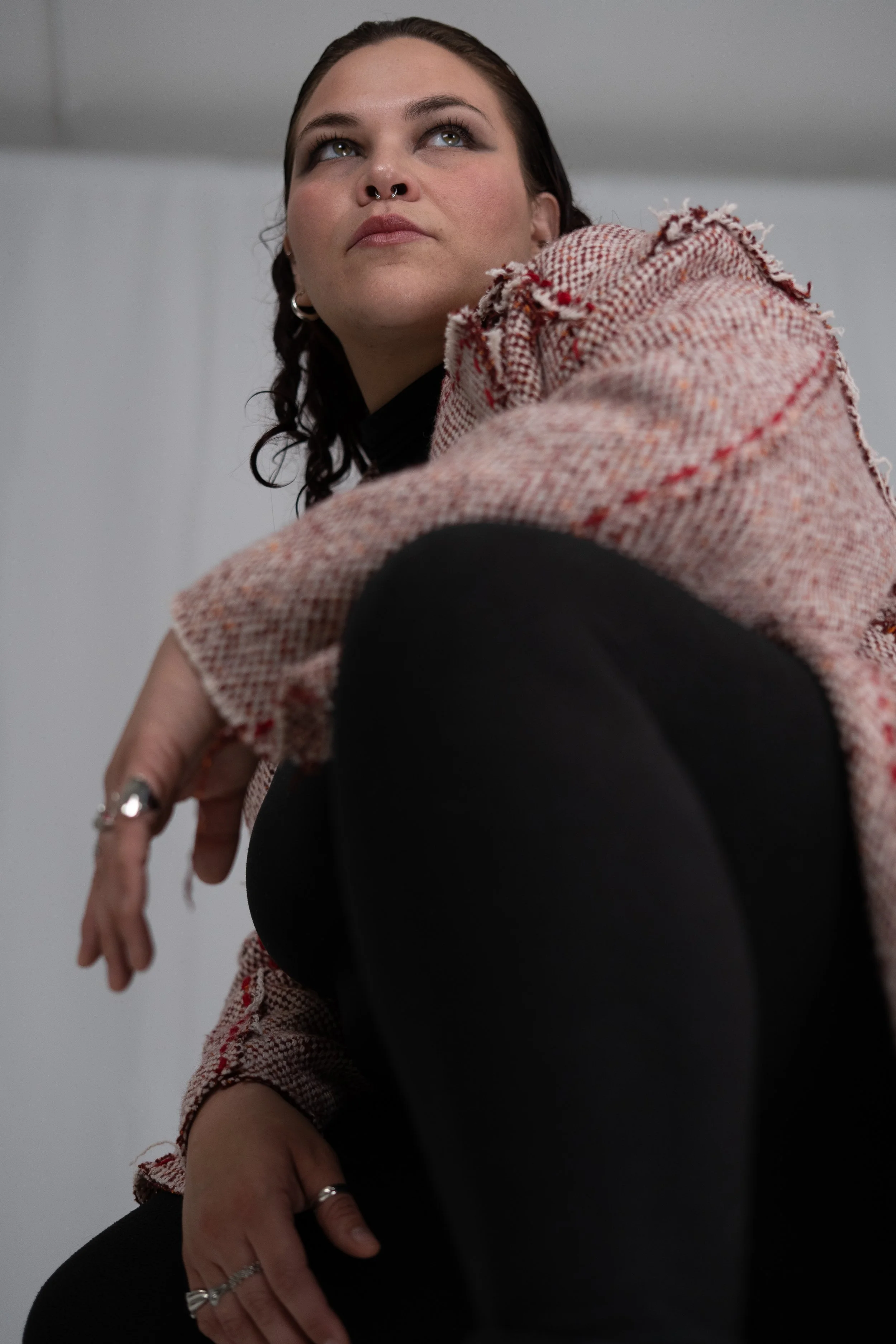 A woman with dark hair styled in braided pigtails looking up, wearing a pinkish tweed blazer over a black top, and multiple rings on her fingers.