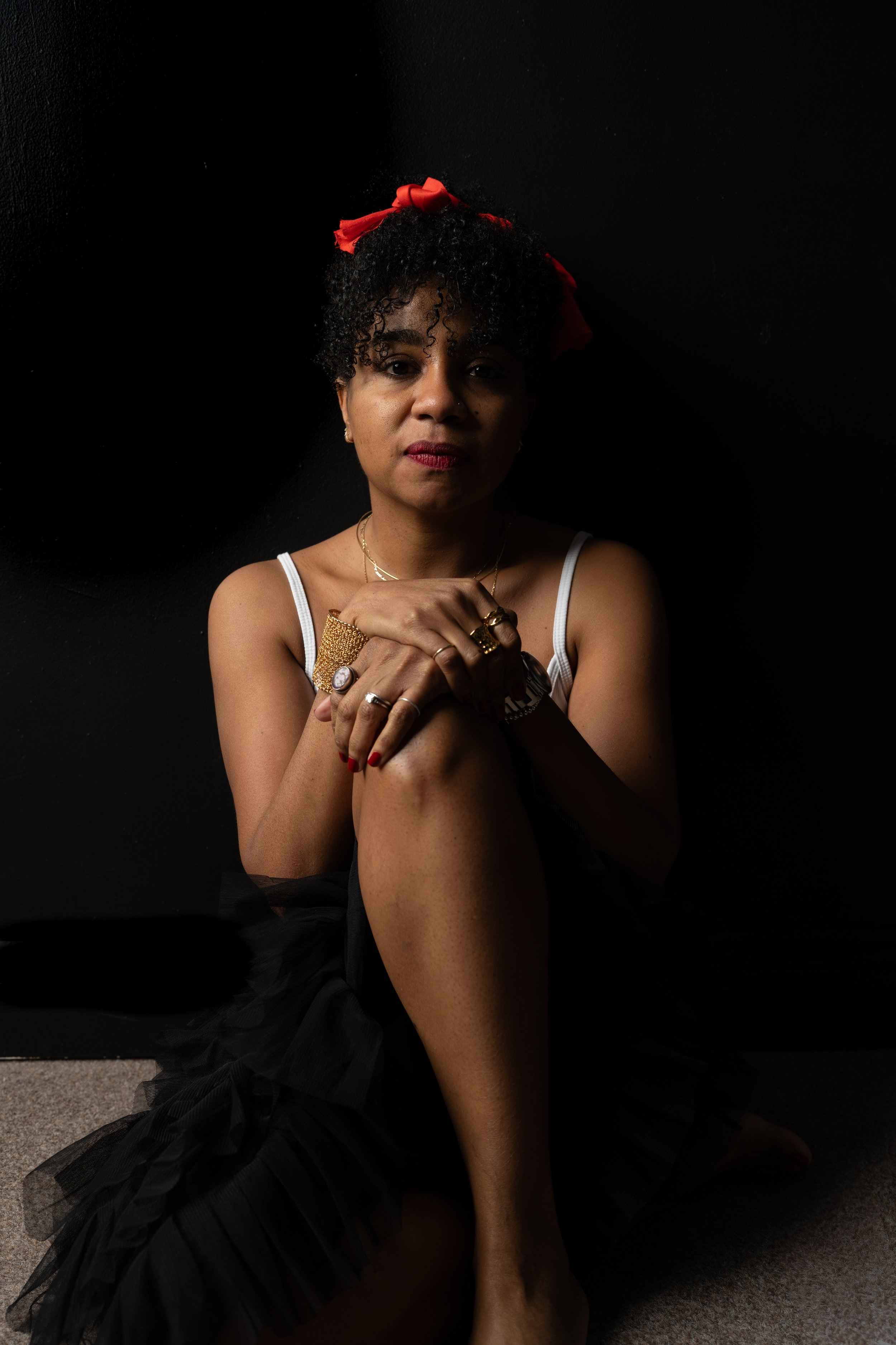 A woman with curly hair wearing a red headband, white tank top, and black skirt, sitting on the floor against a black background.