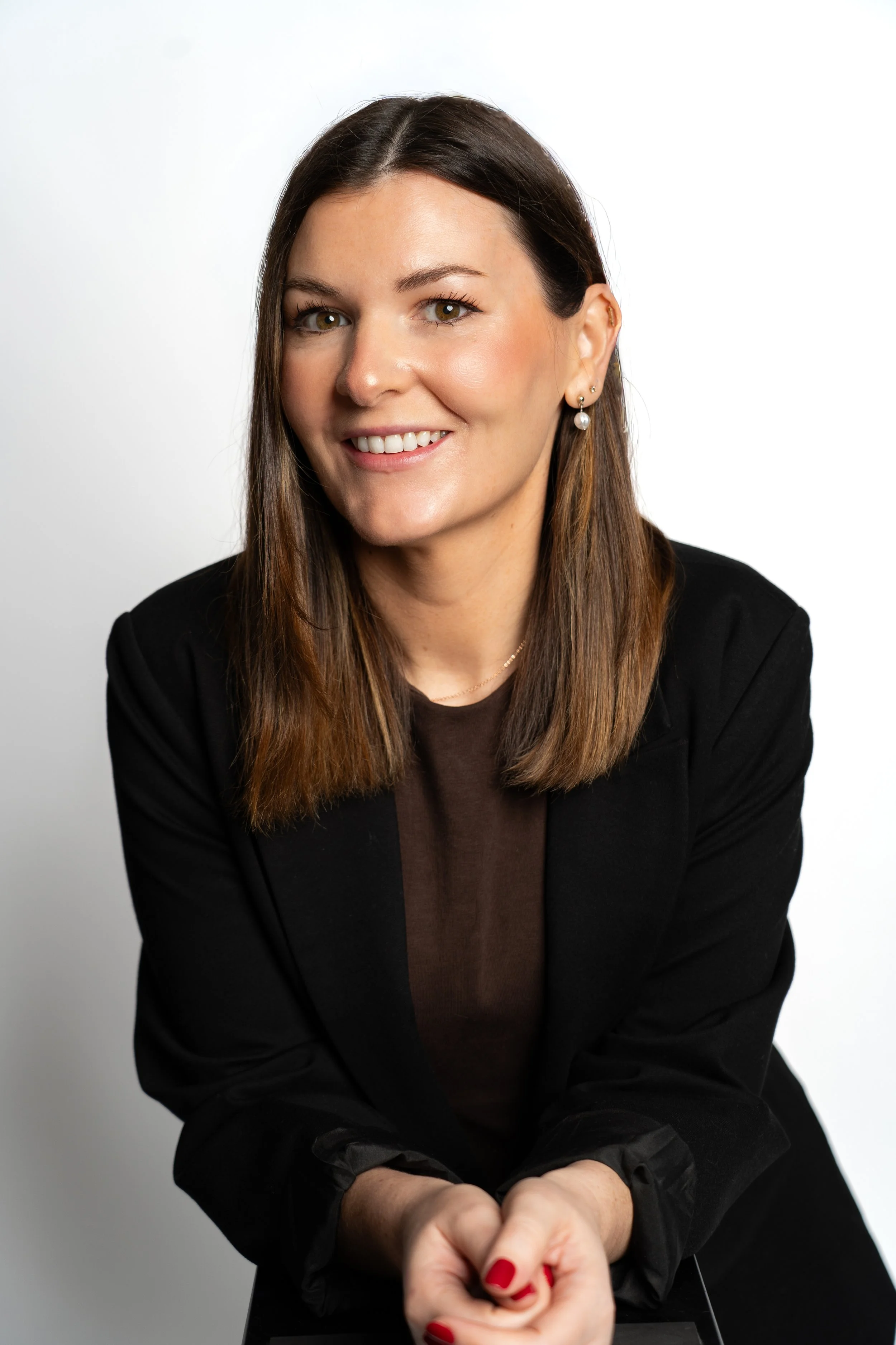 A woman with brown hair, wearing a black blazer and earrings, smiling at the camera against a white background.