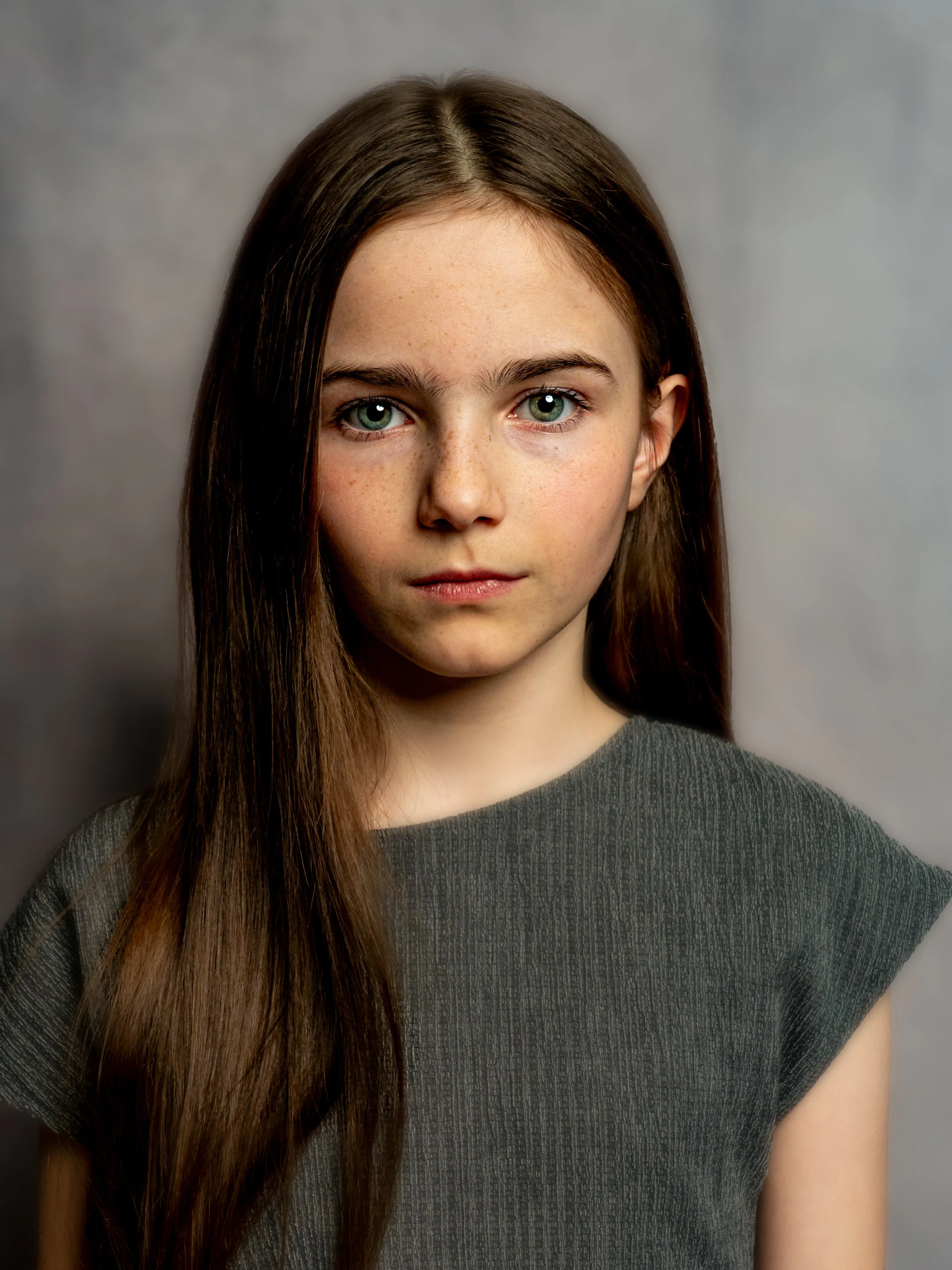 Portrait of a young woman with long brown hair and blue eyes, wearing a dark gray top, against a gray background.