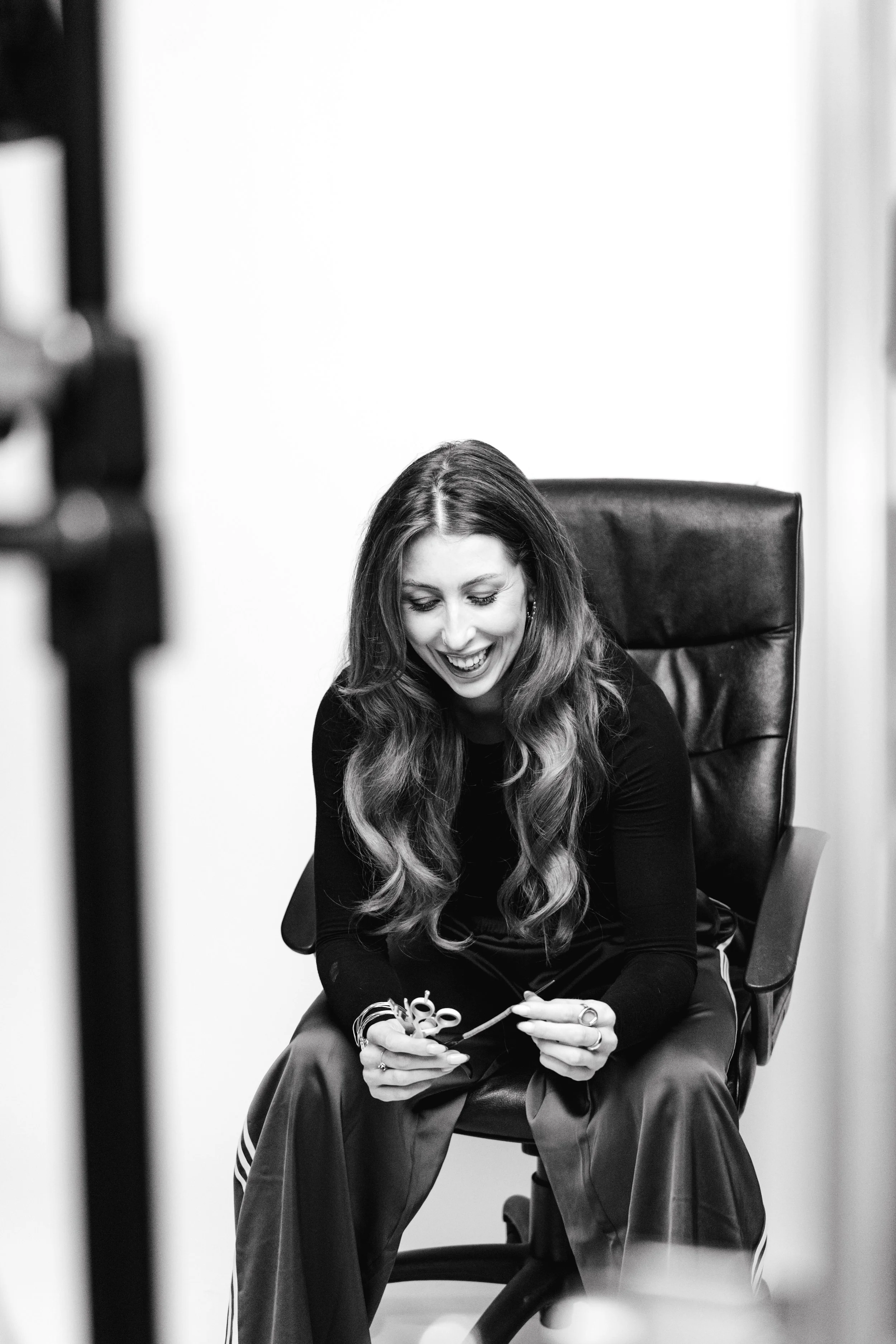 A woman with long hair smiling and looking down, sitting in a black leather office chair, holding scissors and a small object in her hands.