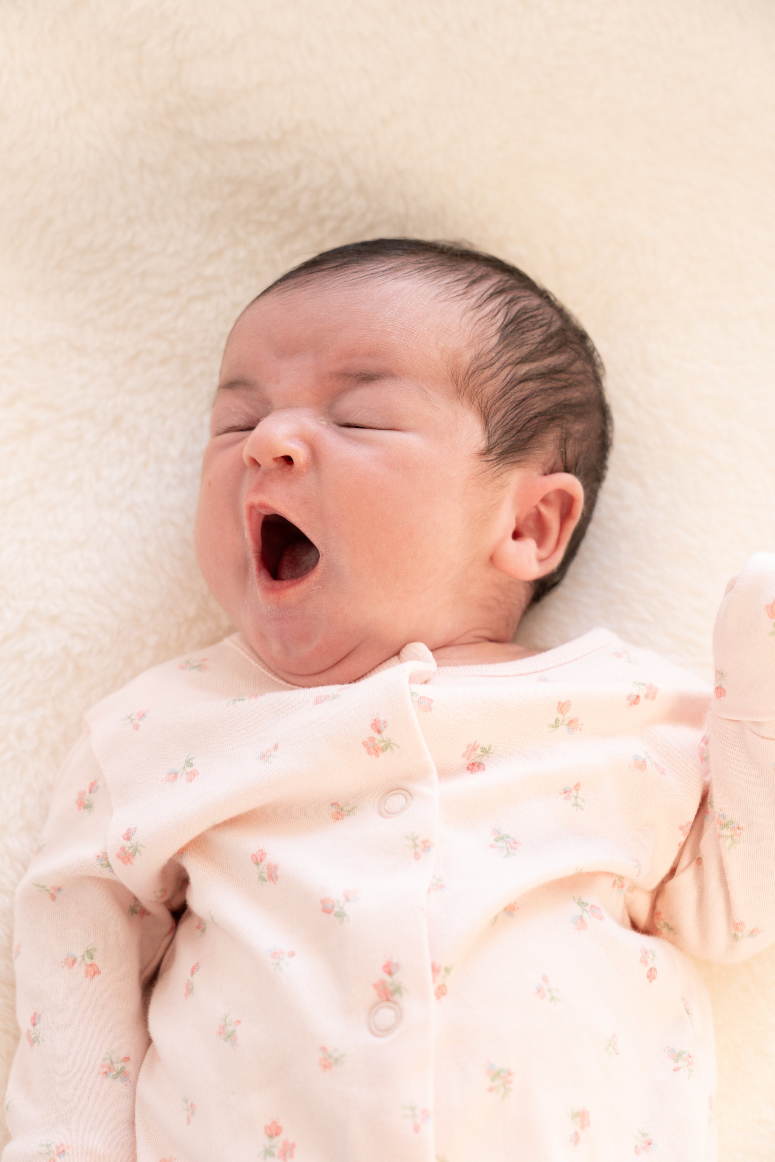 A yawning baby lying on a soft blanket, wearing a cream-colored onesie with small pink and green floral patterns.