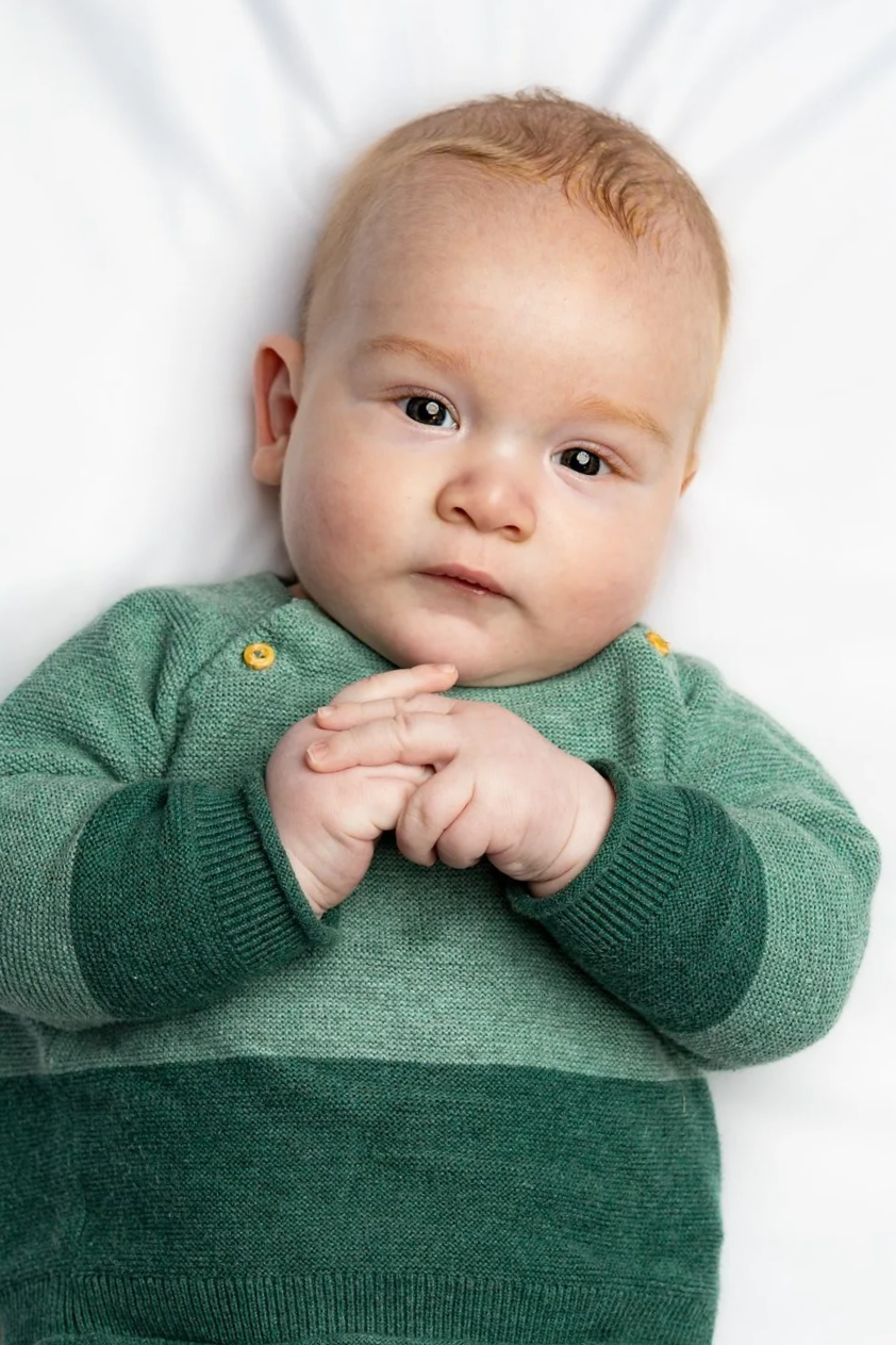 Close-up of a young red-haired baby lying down, wearing a green sweater, with hands clasped together and looking directly at the camera.