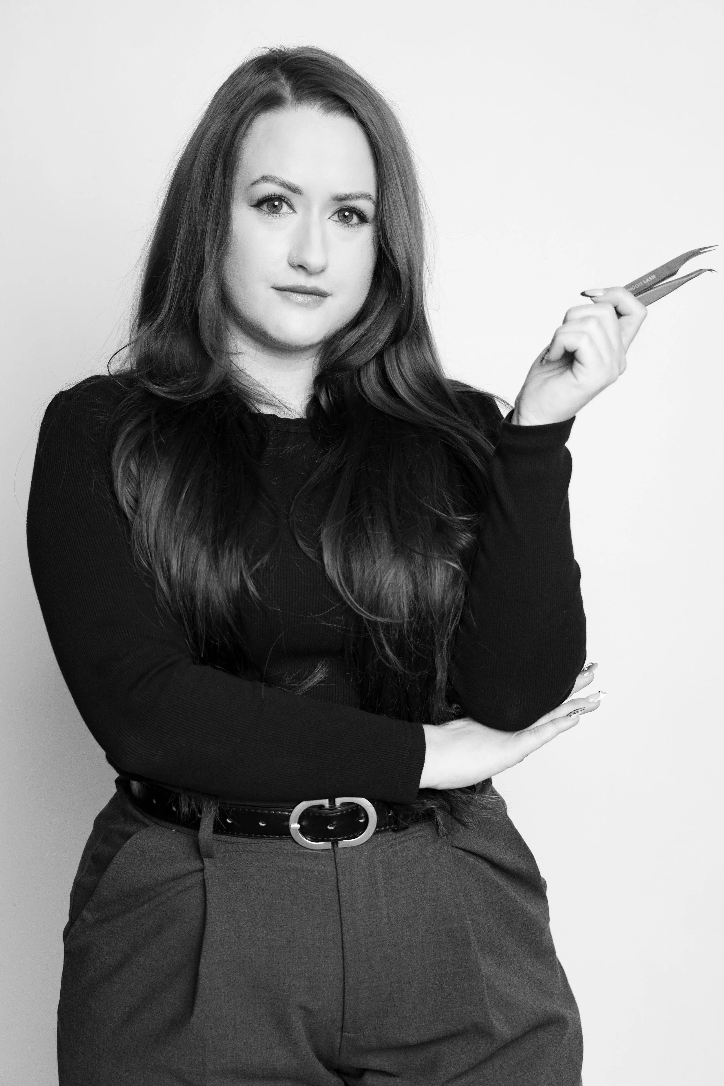 A woman with long dark hair holding a pair of cuticle nippers, wearing a black long-sleeve top and gray pants, standing against a plain background.