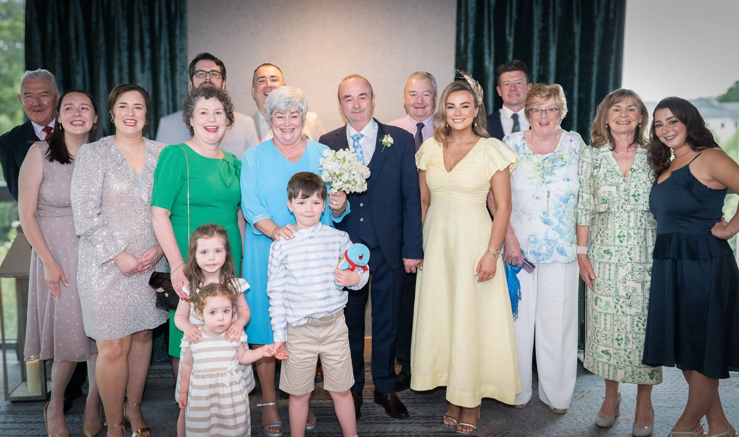 A group of people at a wedding celebration, including the bride, groom, children, and family members, all smiling in a banquet hall with large windows.