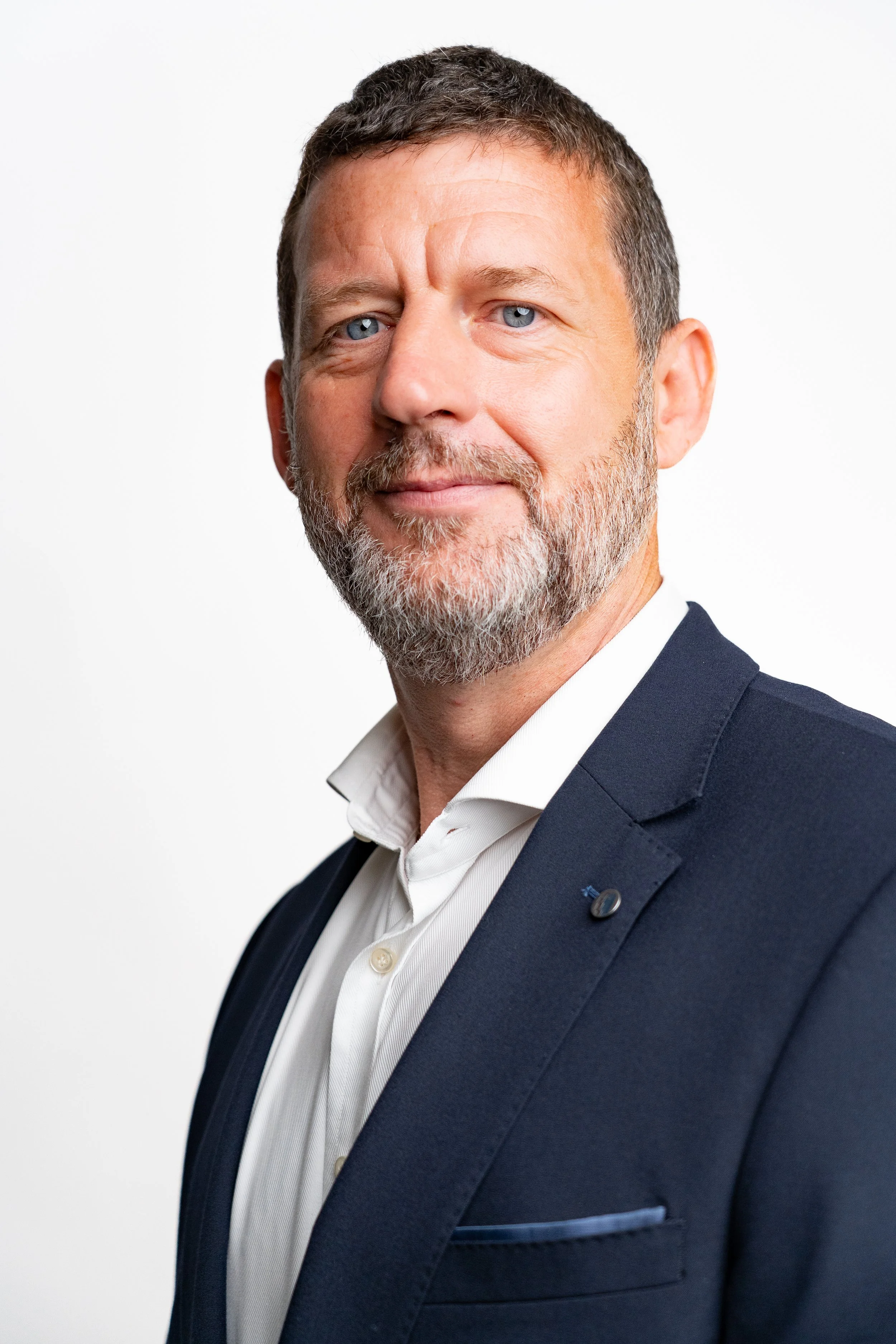 Headshot of a middle-aged man with short brown hair and a beard, wearing a dark suit and white shirt, standing against a plain white background.