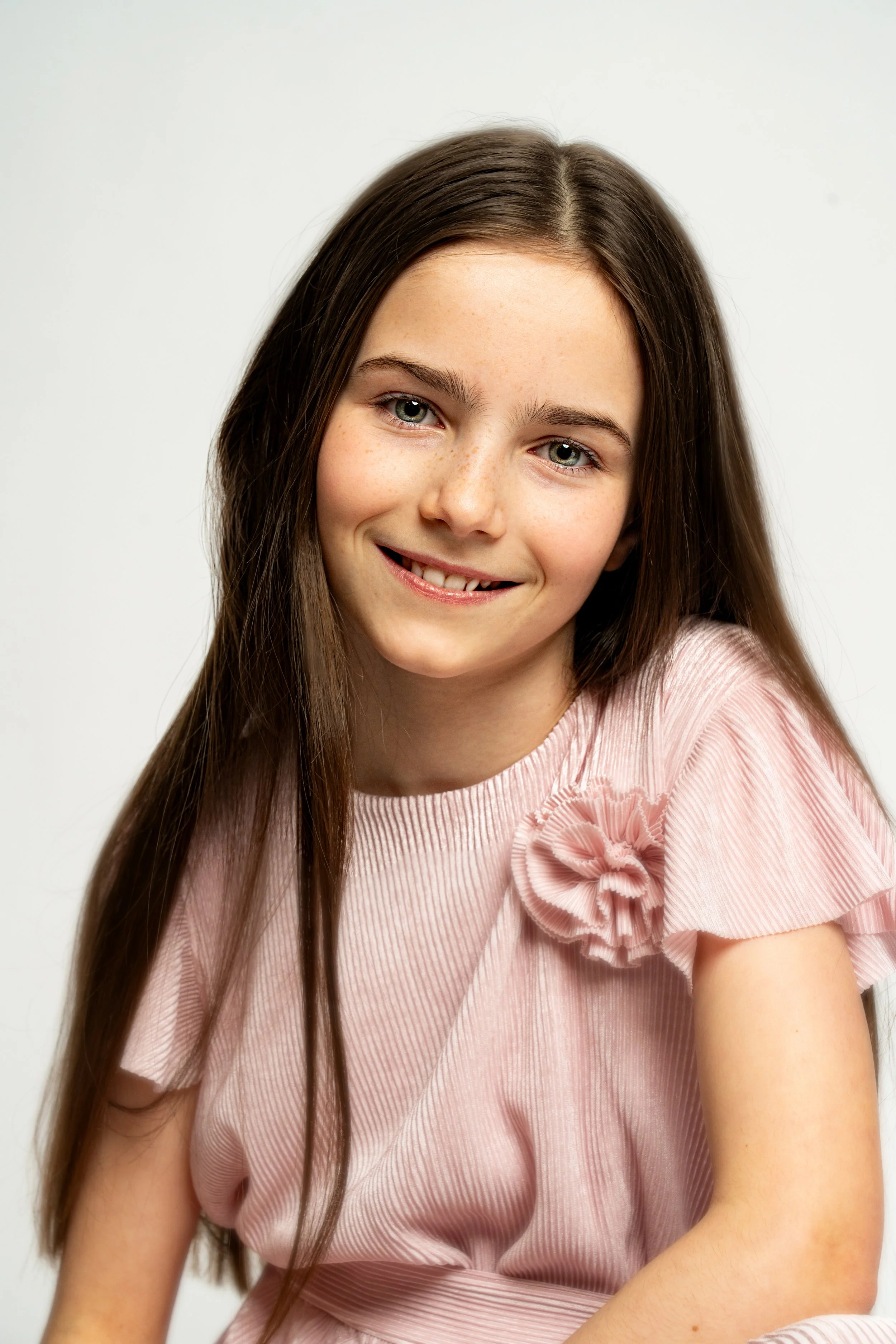 A young girl with long brown hair and blue eyes, smiling, wearing a pink dress with a ruffle flower detail on the shoulder, against a plain white background.