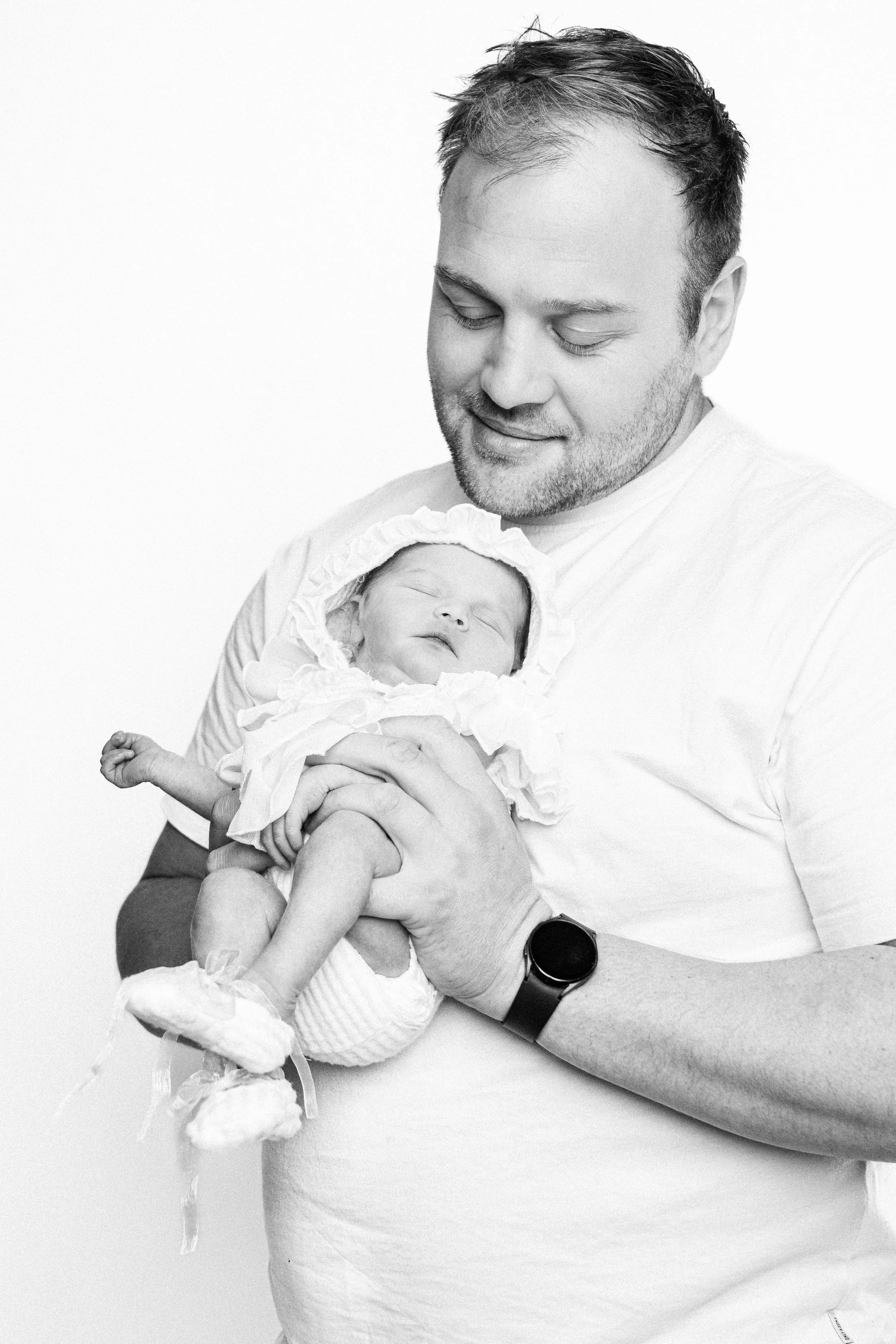 A man holding a newborn baby in his arms, both facing the camera. The baby appears to be sleeping and is dressed in a ruffled outfit with a bonnet. The man is smiling gently and wearing a casual t-shirt and a smartwatch.