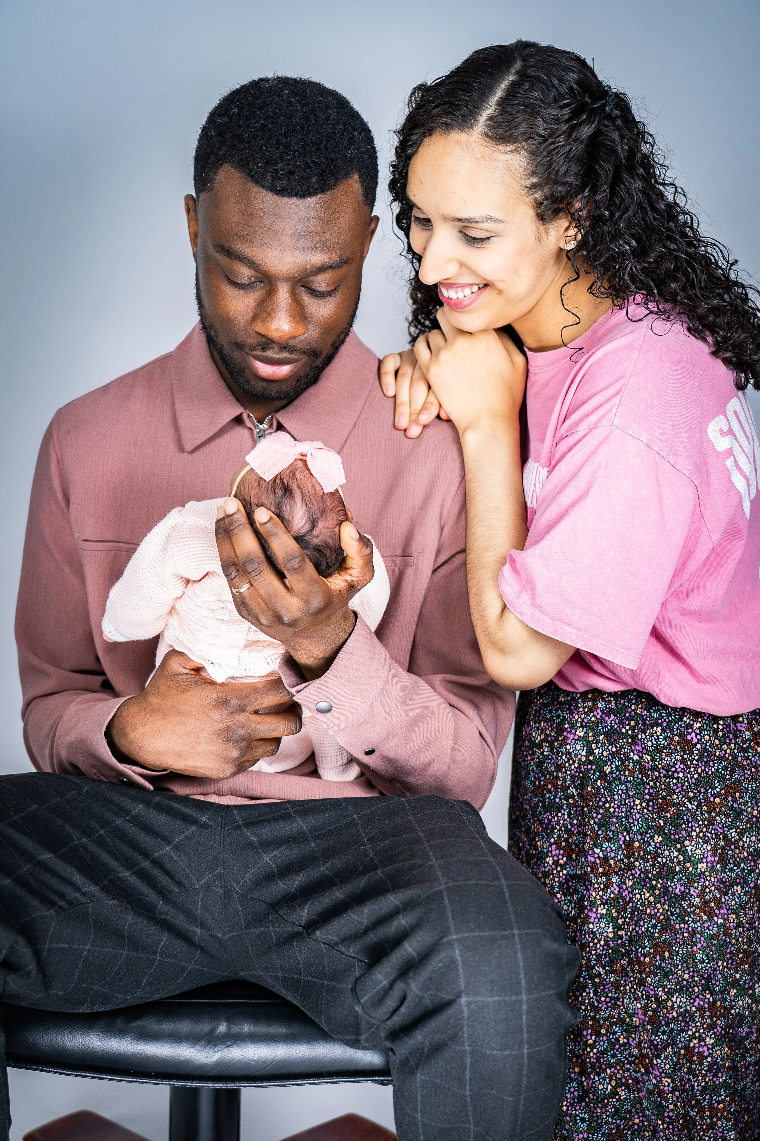 A man in a pink shirt holding a newborn baby, with a woman in a pink t-shirt leaning over and looking at the baby, in a close and happy moment.