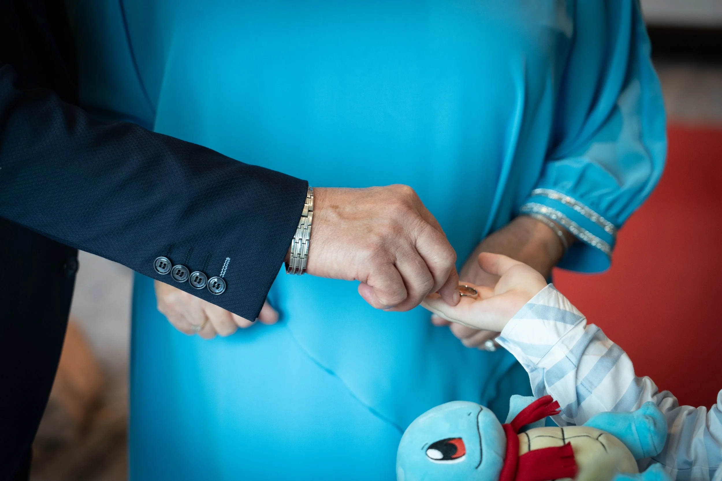 A person in a dark suit is handing a ring to a child, who is sitting in a blue chair with a toy doll on their lap. An adult woman in a blue dress observes the exchange.