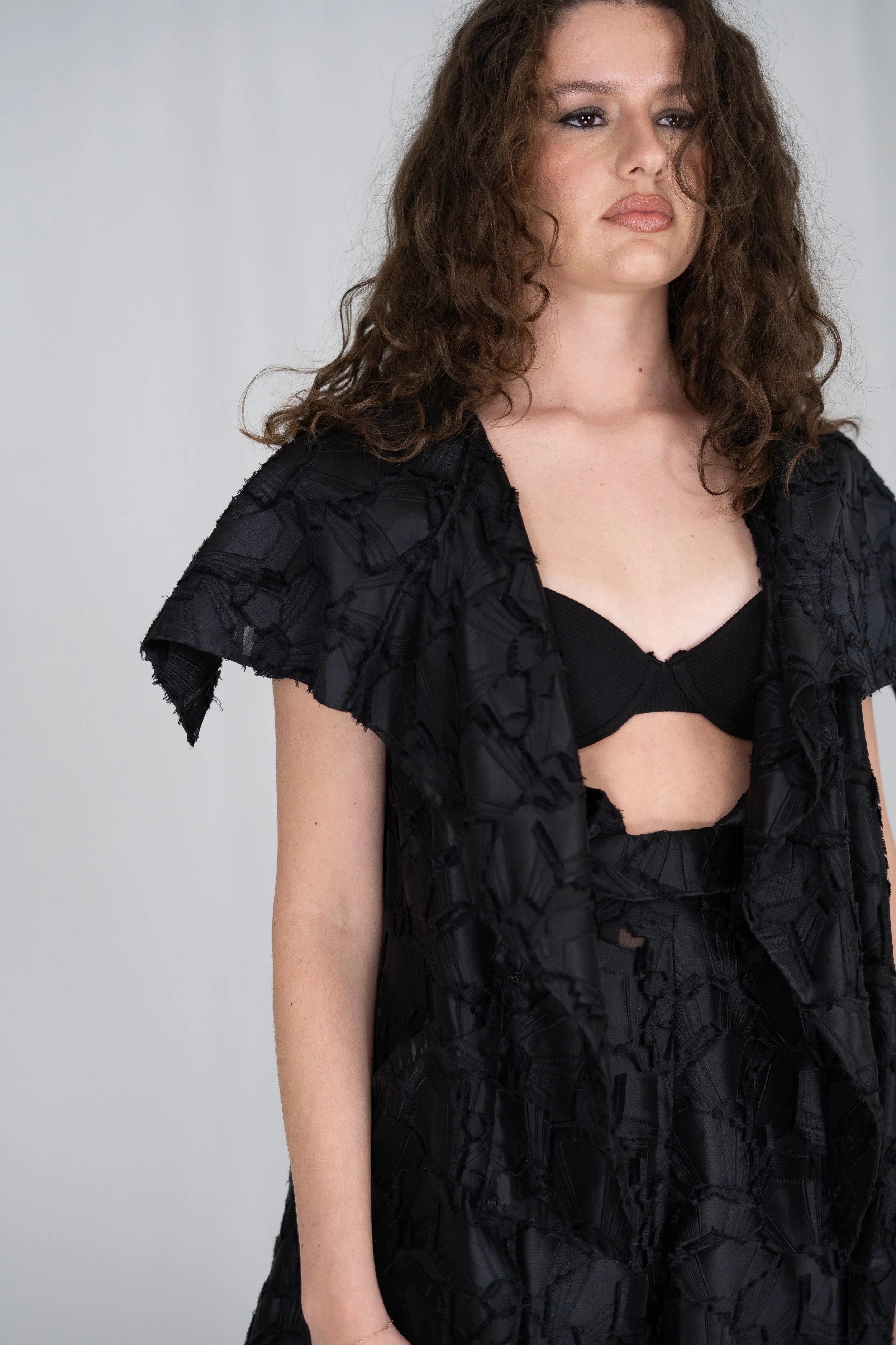 A woman with curly brown hair wearing a black textured top and a matching black textured bra, standing against a plain light gray background.