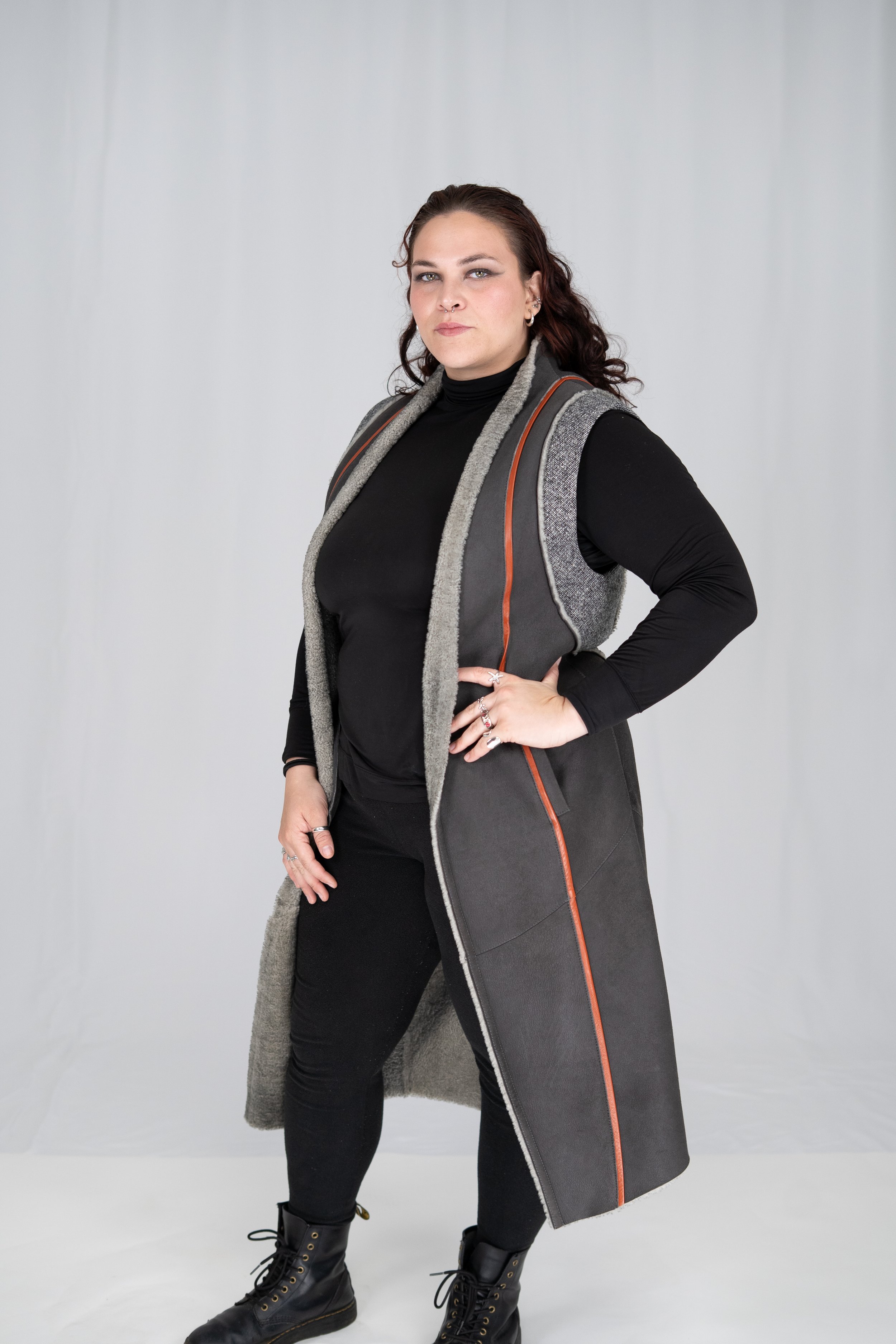 A woman with wavy brown hair, dressed in all black with rings, stands confidently with her hand on her hip, wearing a sleeveless coat over a black turtleneck, in front of a plain gray background.
